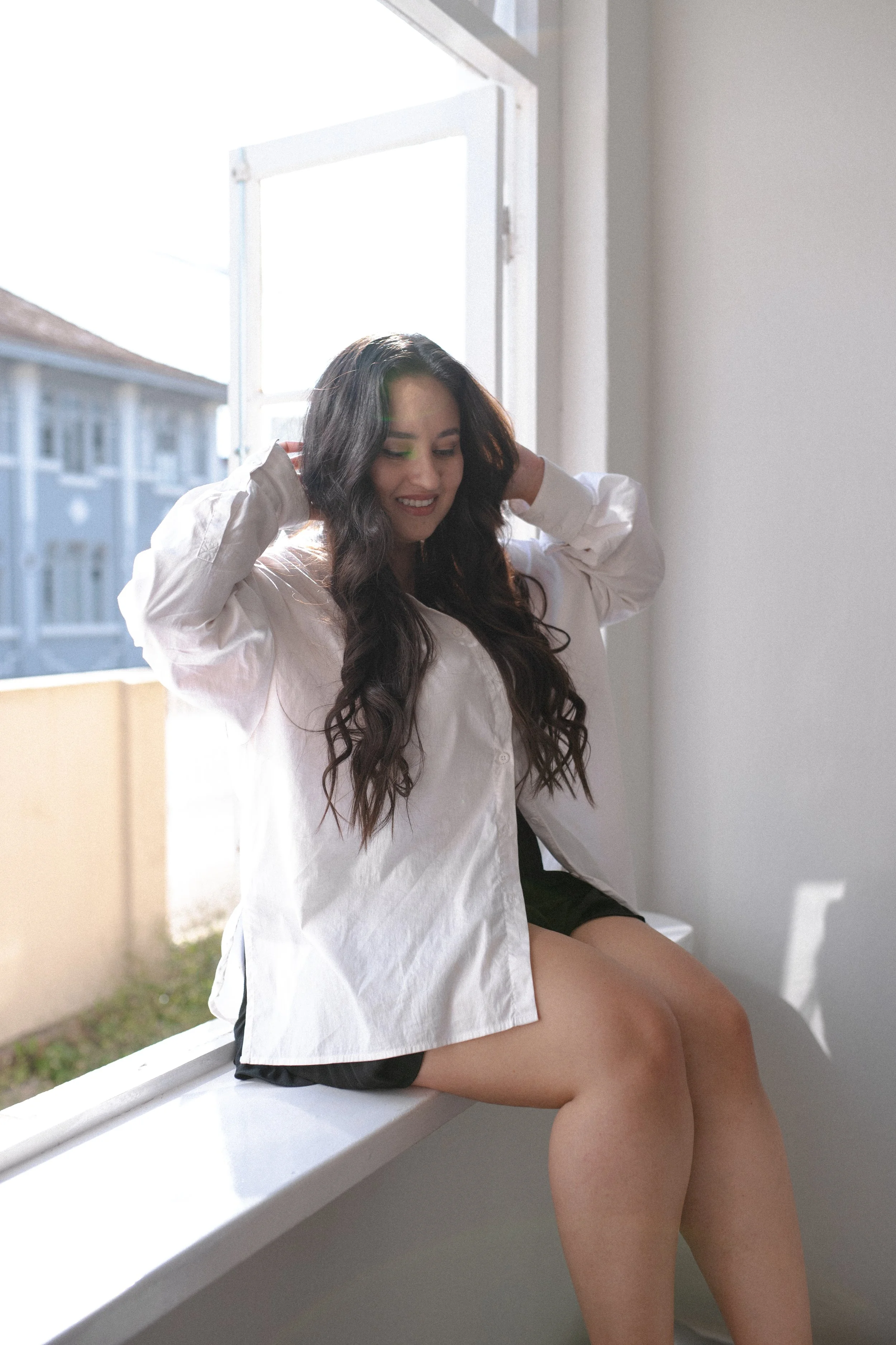 Mulher com cabelo longo e escuro sorrindo, sentada na janela, vestindo camisa branca e shorts pretos, com luz do sol entrando pela janela ao fundo.