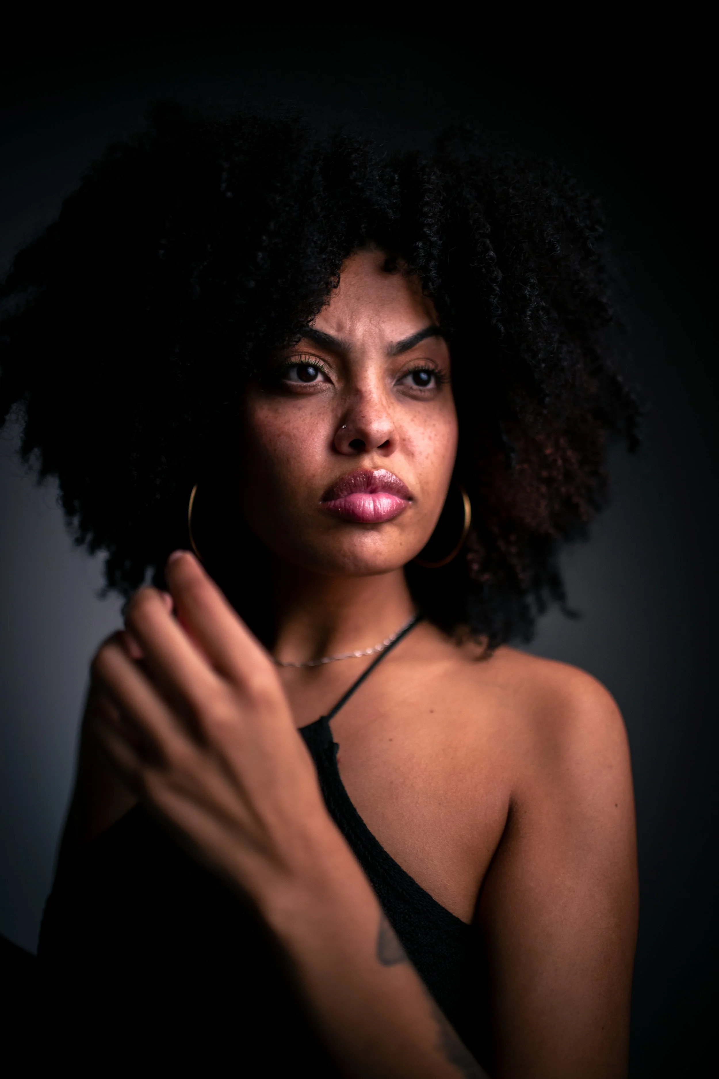  retrato de uma mulher negra com cabelo cacheado escuro, usando brincos de argola dourados e camiseta preta, olhando seriamente para a câmera, com fundo escuro.
