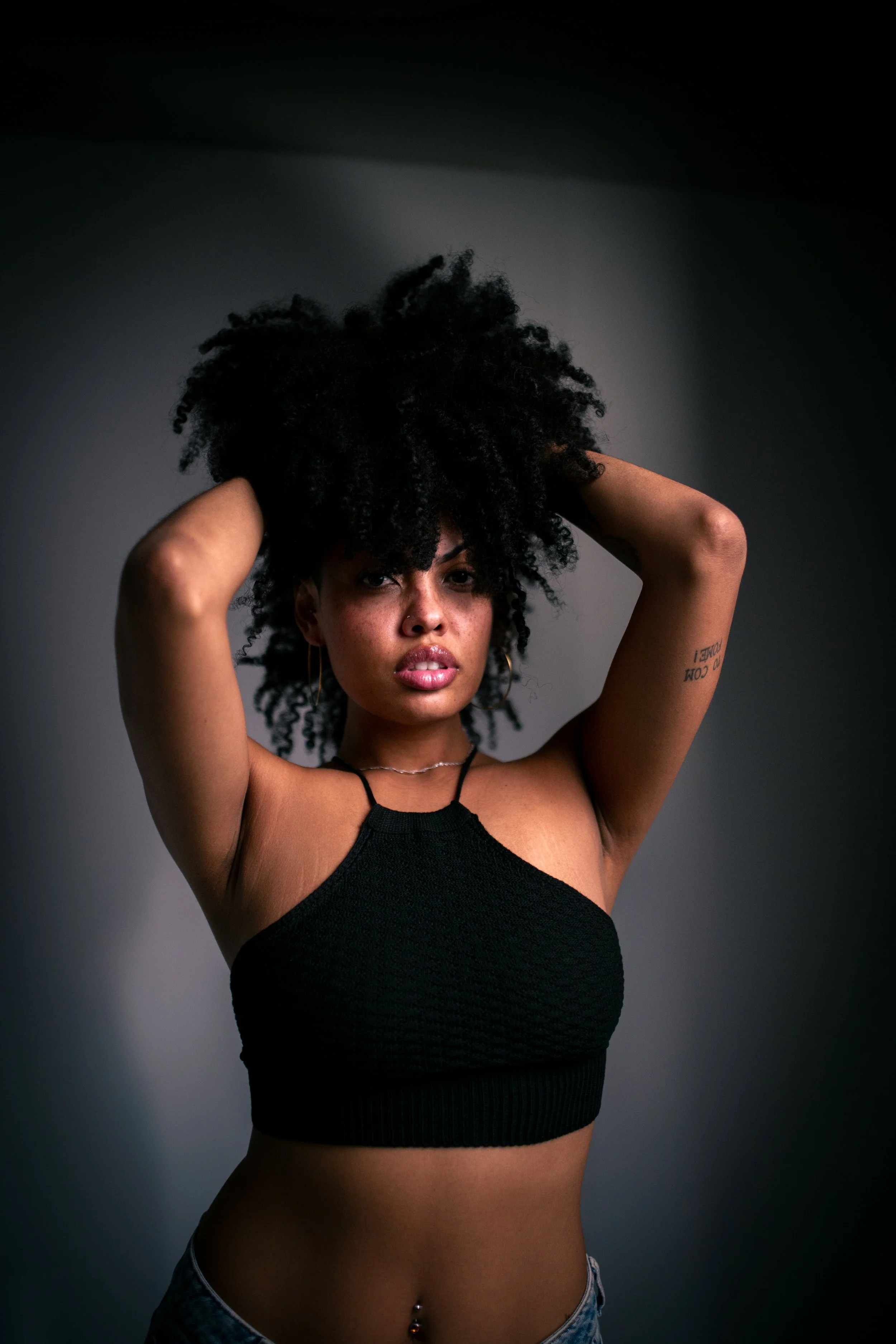 Mulher com cabelos cacheados pretos, vestindo top preto e jeans, olhando seriamente para a câmera, com braço levantado e expressão confiante.