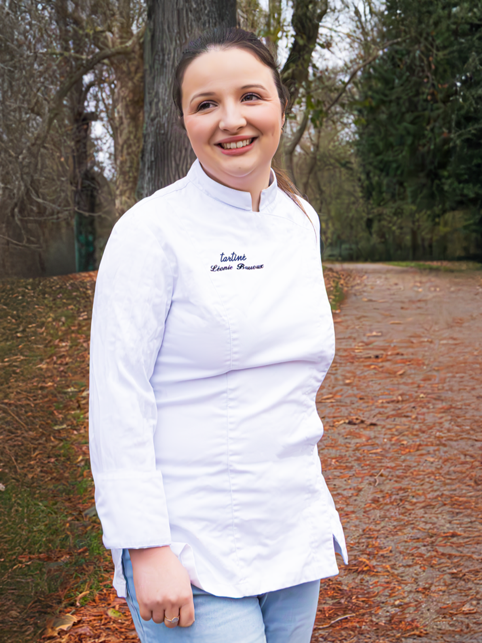 Jeune femme souriante, habillée d'une blouse blanche de chef, dans un parc en automne, avec des arbres et un chemin en arrière-plan.