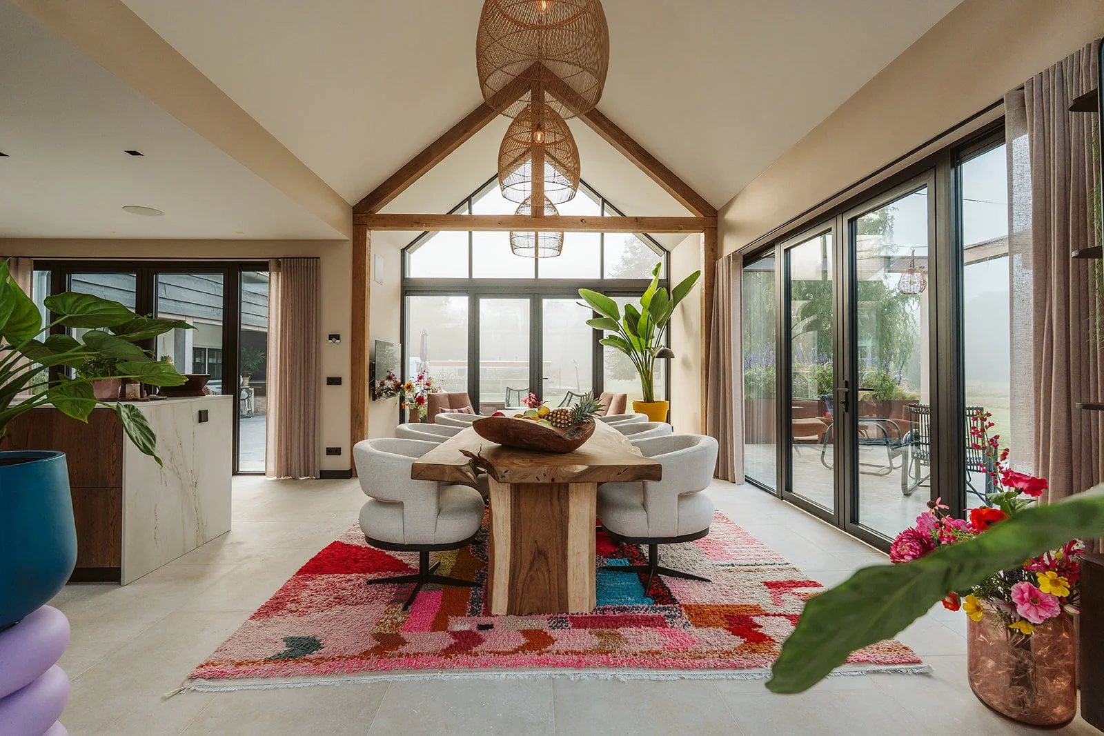 Bright and airy dining room with large glass sliding doors, a wooden dining table with white chairs, colorful rug, large green indoor plant, and decorative pendant lights.