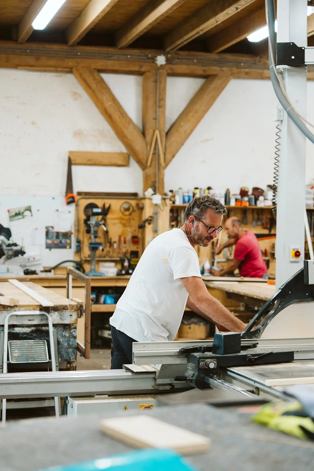 Two men working in a woodworking shop; one man is operating a table saw, and the other is working at a workbench in the background.