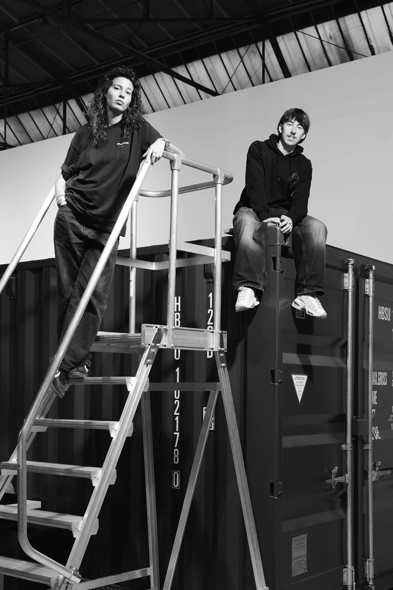 zwei mitarbeiter*innen von 711rent fotoequipment verleih in hamburg posen cool für die kamera, einer auf einem container sitzend, die andere auf einer Leiter