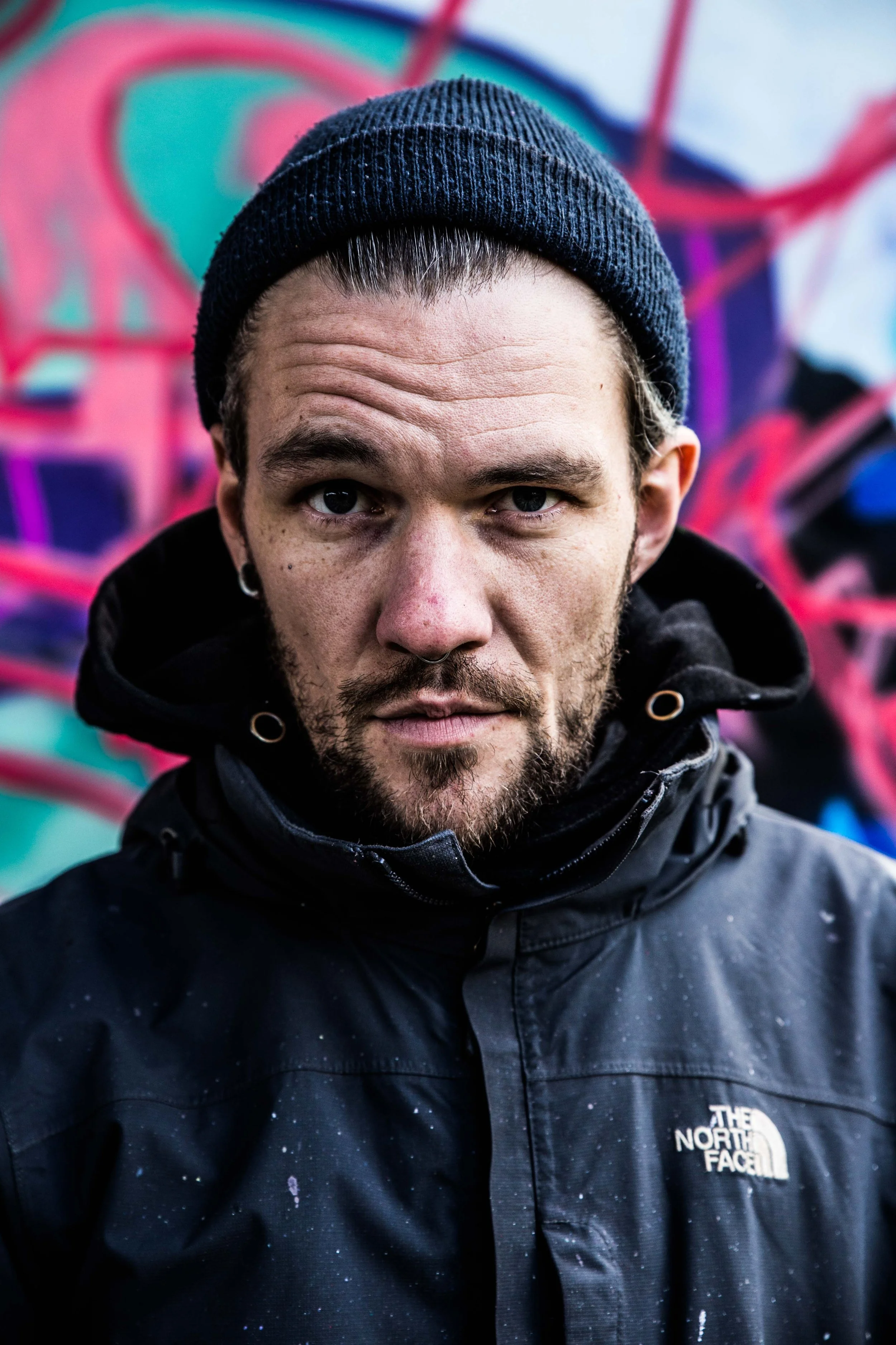Kieler Graffiti Künstler*in Carsten/Caro mit Mütze und in Northface Jacke guckt intensiv in die Kamera vor pink-blauer Graffitiwand
