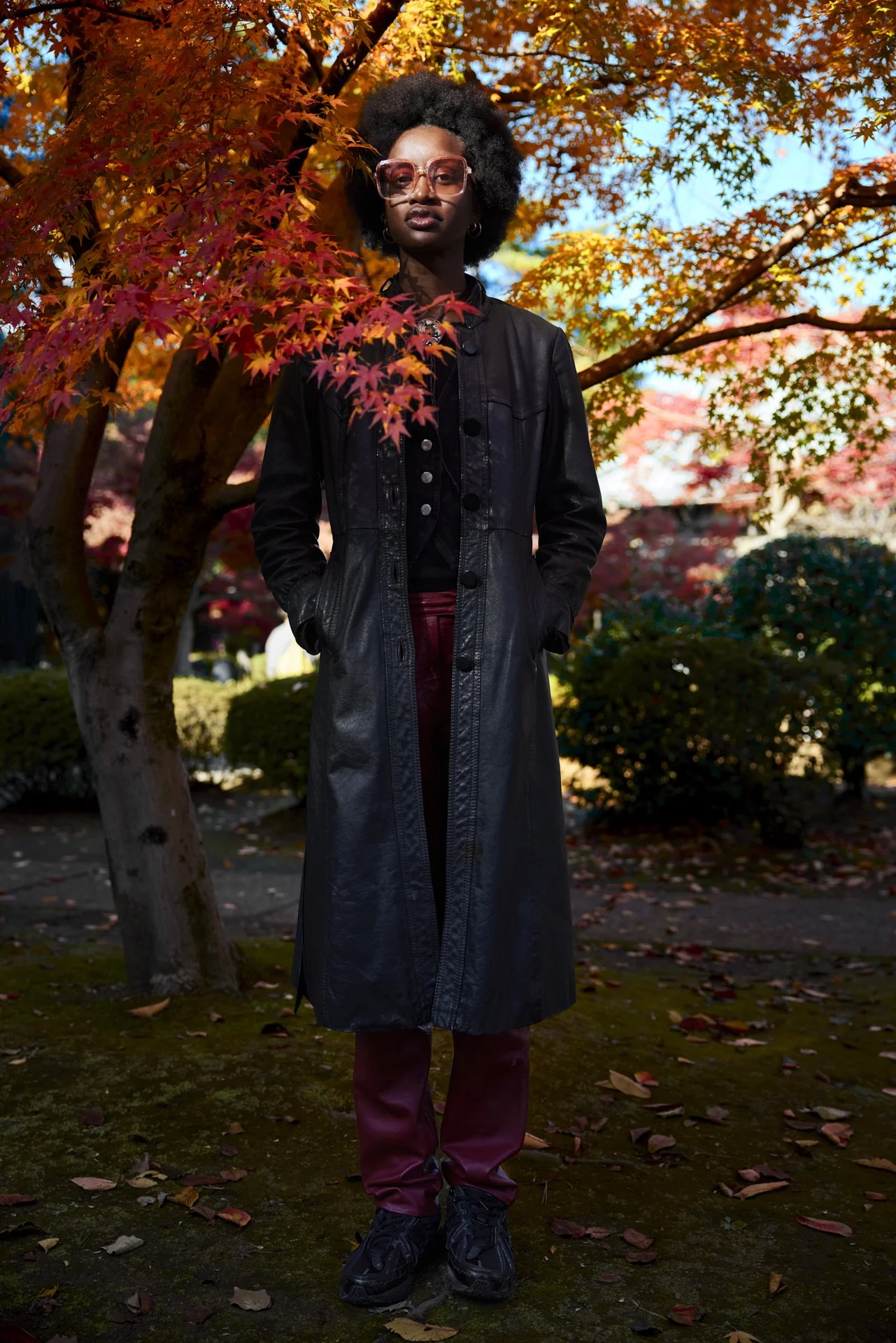 Schwarzes Model steht in rot blühendem Ahornbaum in Tokio, Japan mit Sonnebrille auf und langem schwarzem mantel