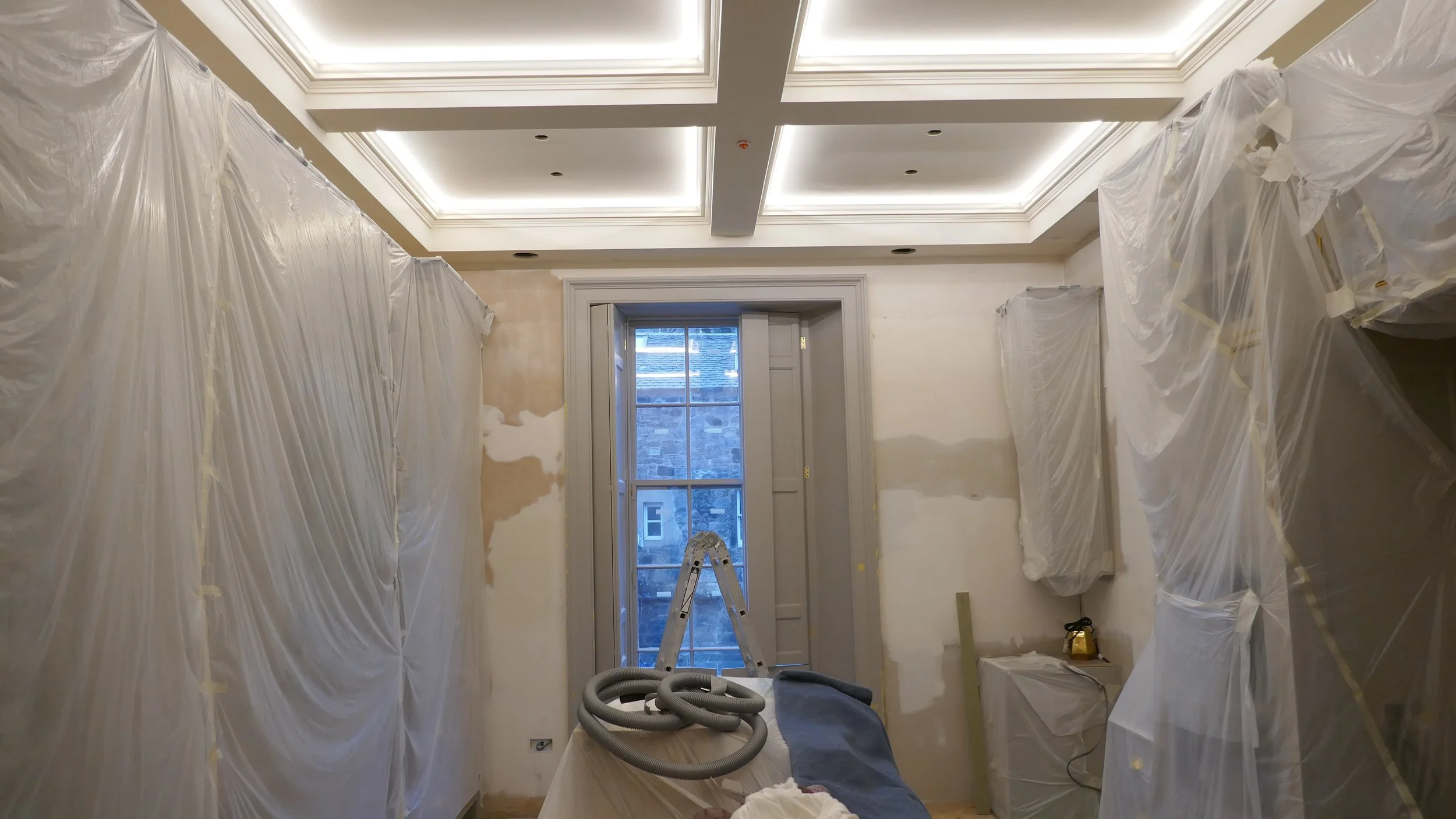 Interior room under renovation with plastic coverings on the walls, a ladder by the window, a vacuum hose on a wrapped table, and construction tools surrounding.