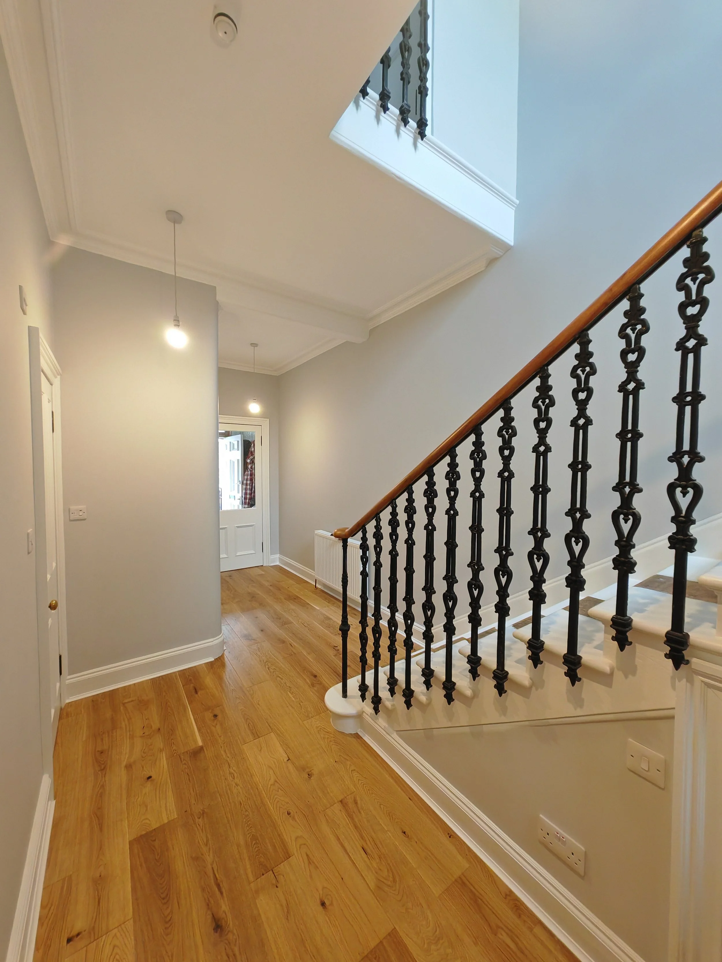 Interior view of a hallway with wooden flooring, a staircase with ornate black iron balusters, and a staircase with a wooden handrail. The walls are painted in a light color, and there are several small hanging ceiling lights.