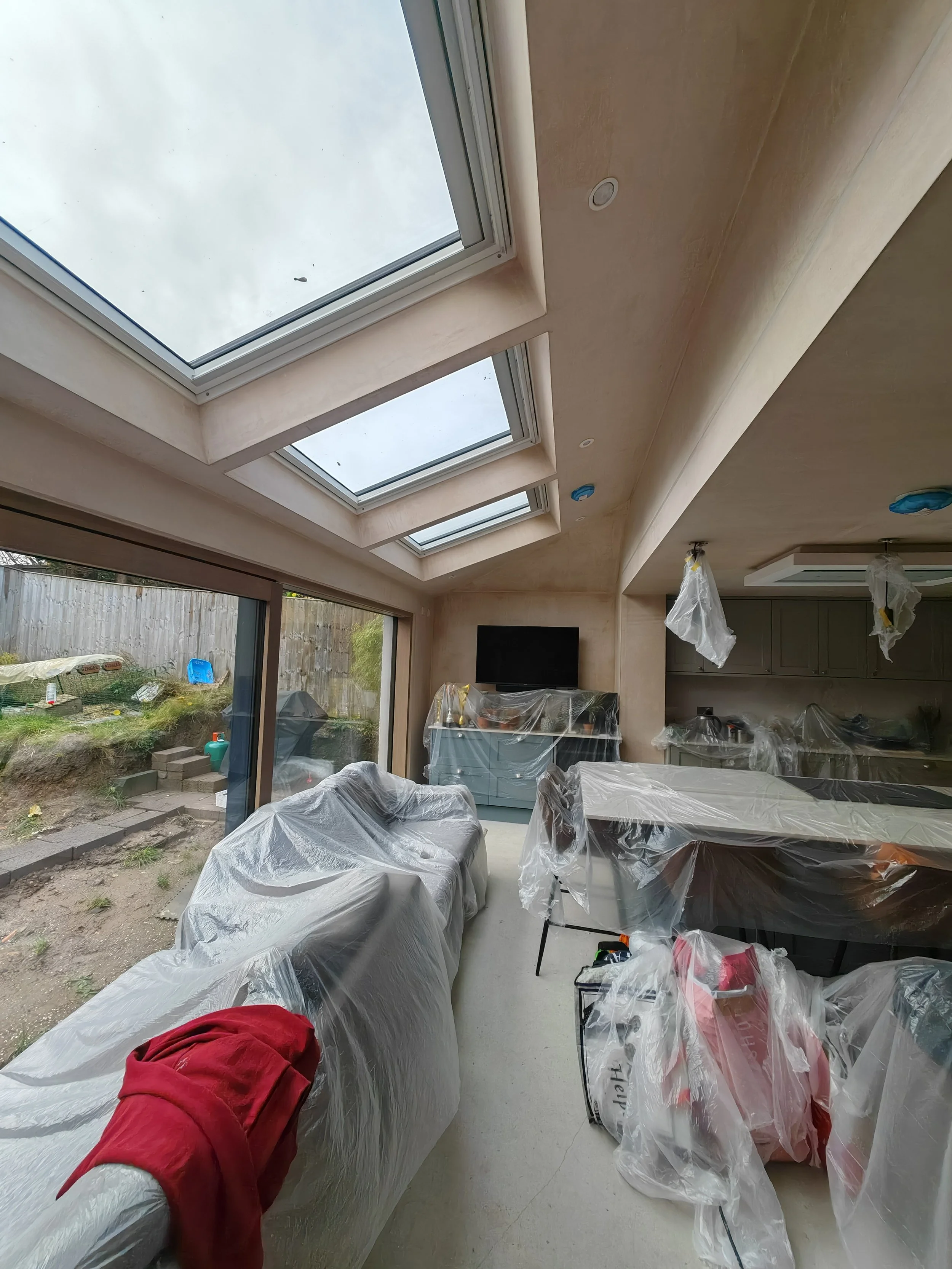 A living room with large skylights and a sliding glass door leading outside, furniture and kitchen area covered in plastic for renovation or painting.