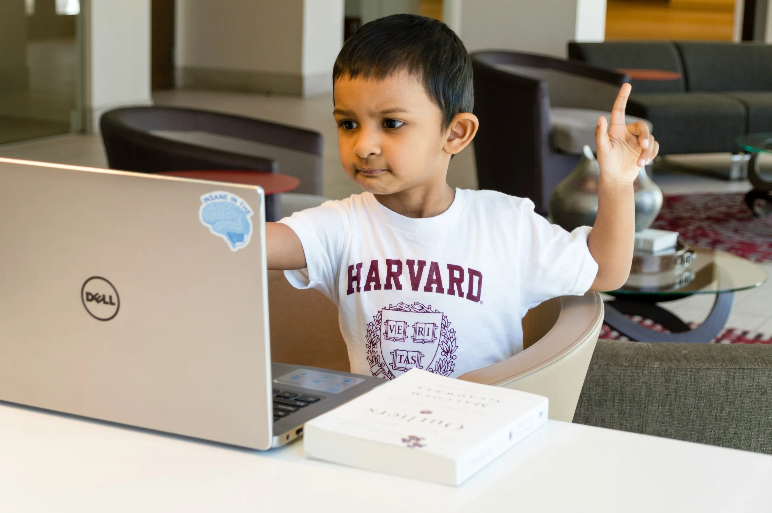 A young boy sitting at a table, looking at a laptop. He is wearing a Harvard T-shirt and has one hand raised with his index finger pointing up.