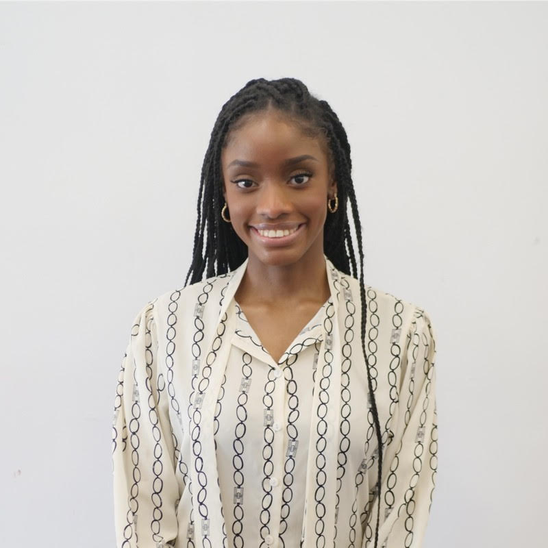 A young woman with long braided hair, wearing a cream-colored blouse with black chain patterns, smiling and standing against a plain white wall.