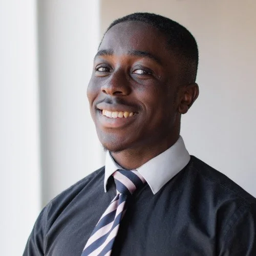 A young man in a business shirt and striped tie smiling near a window.