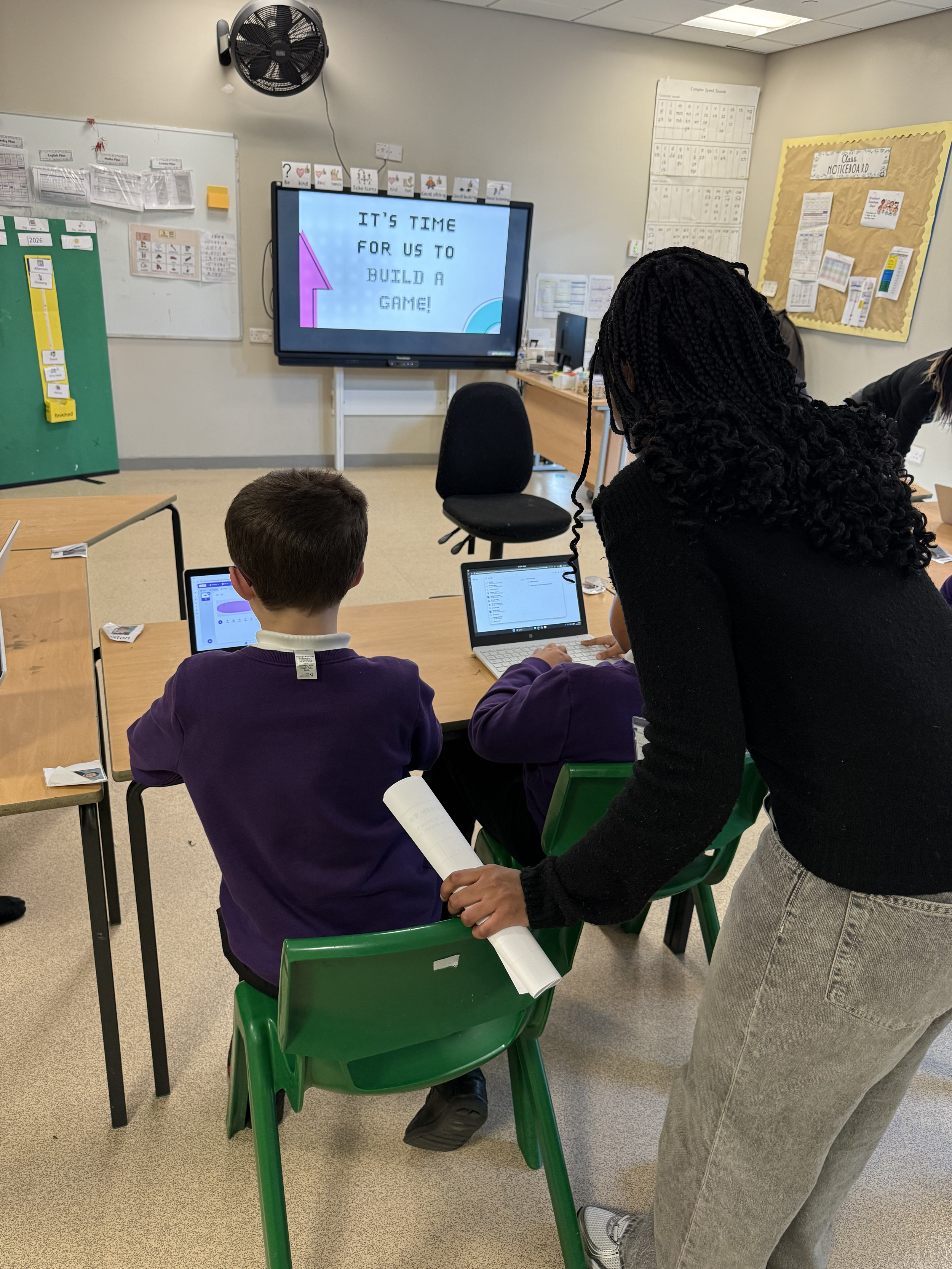 A classroom with students working on laptops. A teacher is leaning over a student to assist. A digital screen in the front displays the message, 'It's time for us to build a game!'