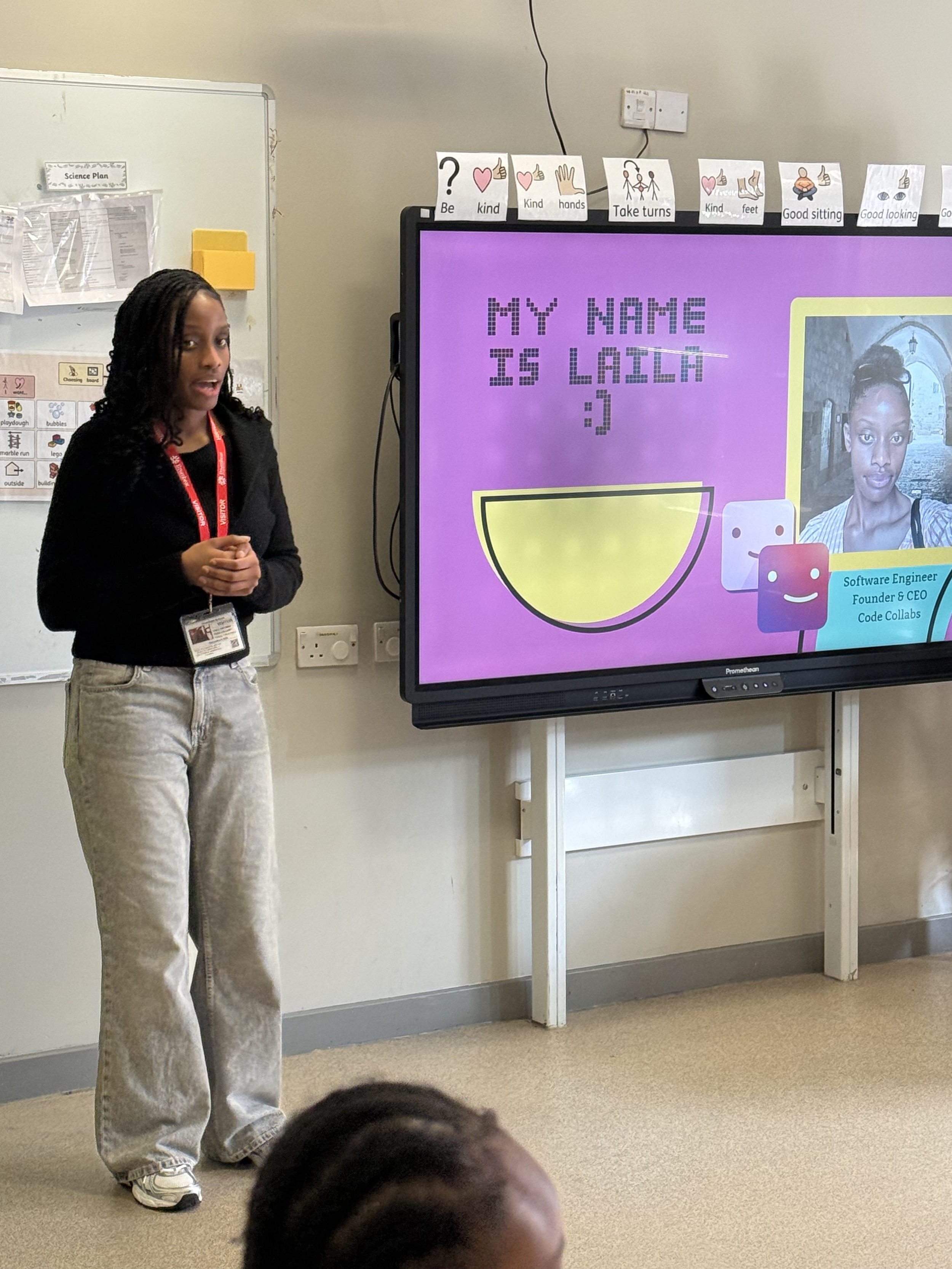 Young girl standing in front of a classroom presentation screen displaying her name and a photo of herself, with a whiteboard and classroom decorations in the background.