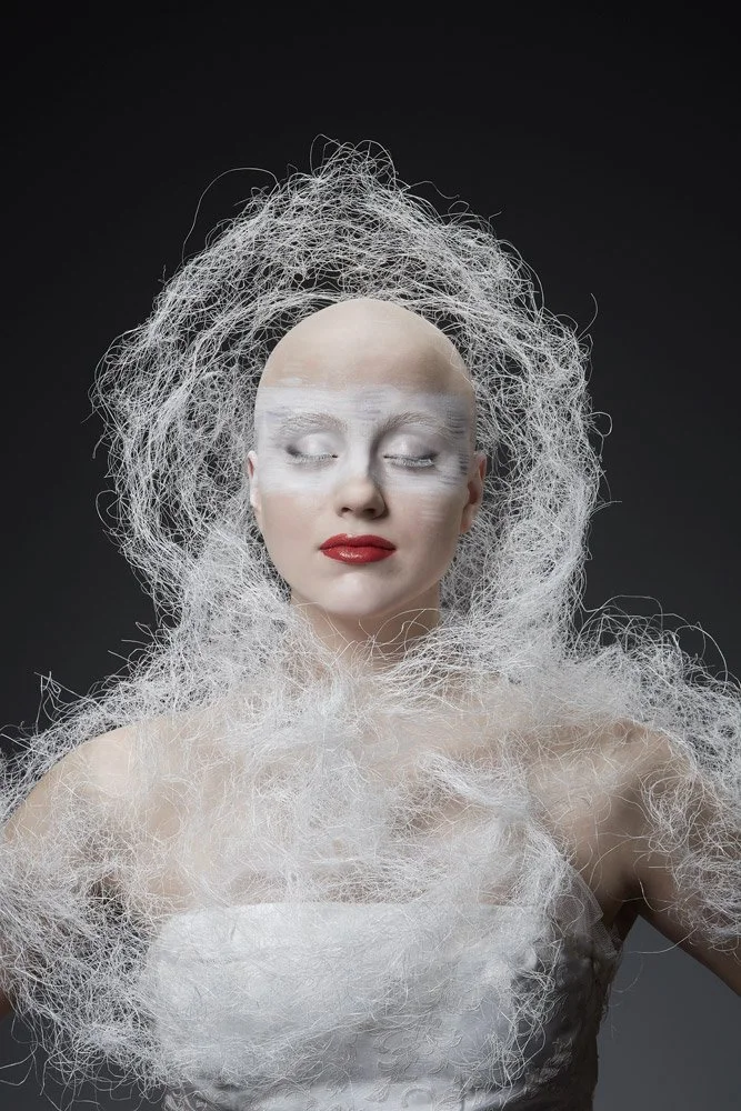 Künstlerische Portraitaufnahme einer Frau mit weißem Make-up, roten Lippen und kunstvoll gestaltetem weißen Haar aus feinem Netz, das um Kopf und Schultern drapiert ist, vor dunklem Hintergrund.