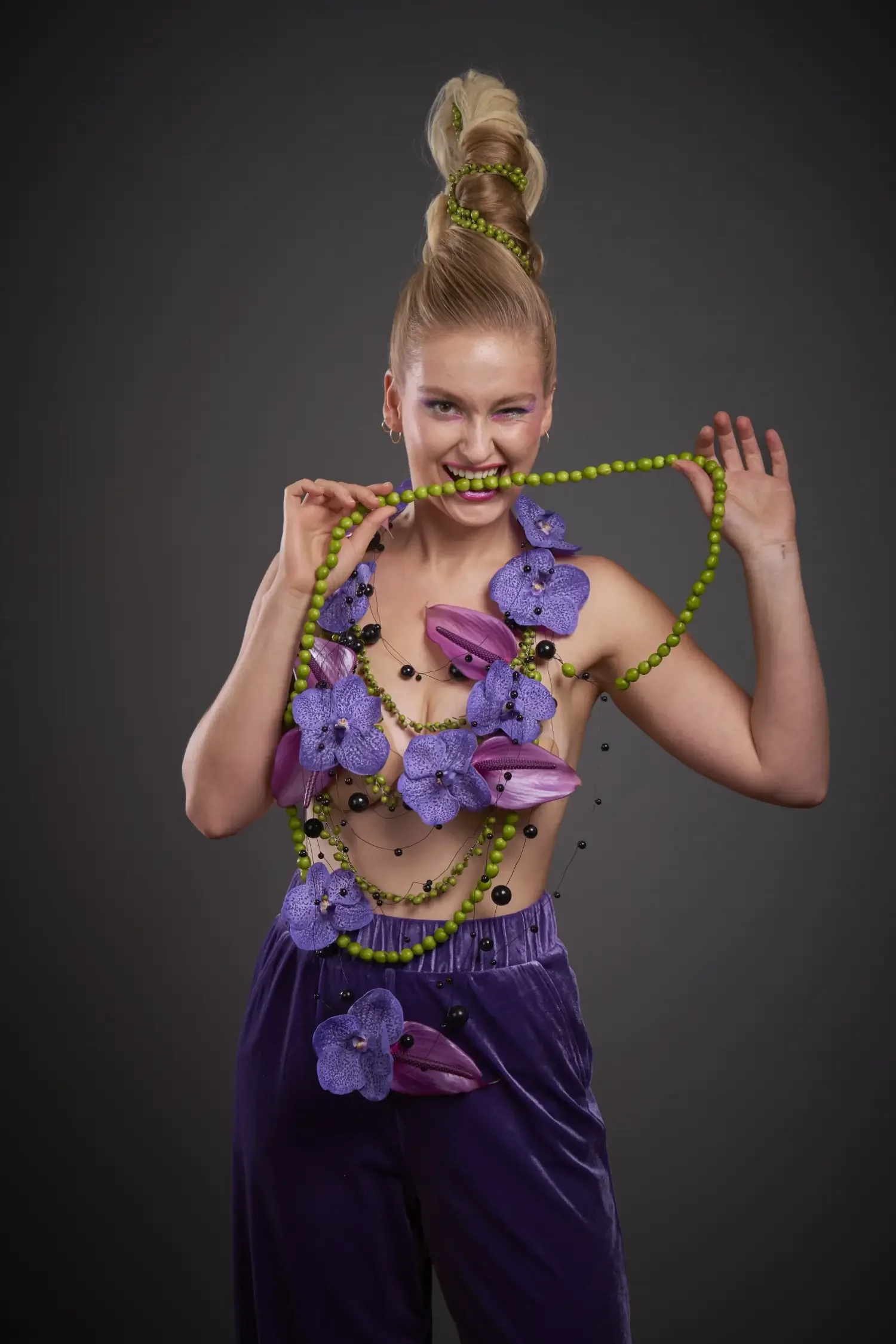 Avantgardistisches Fashion- und Beautyportrait aus der Serie „Botanical Avantgarde“ von Garzareck mit skulpturalem Hochsteck-Hairstyling, grüner Perlenkette, violetten Orchideen und floraler Couture vor dunklem Studiohintergrund.