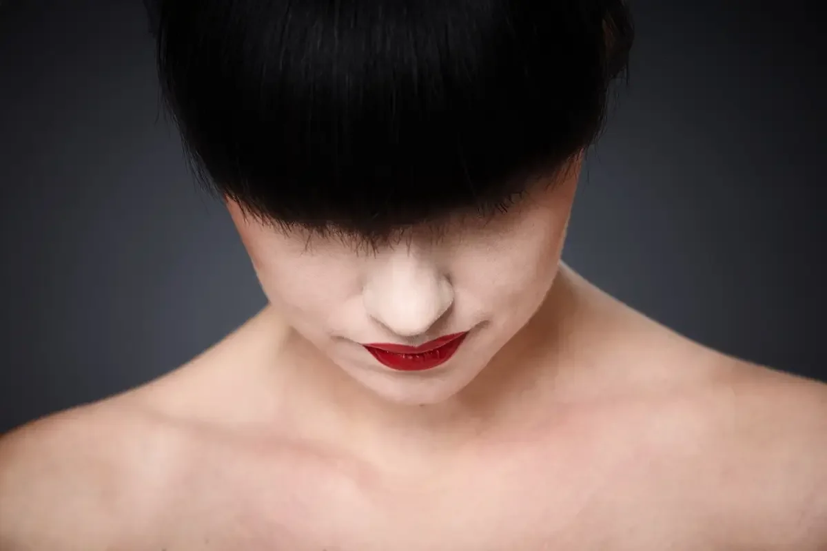 Minimalistisches Close-up eines Beauty-Editorials mit tiefschwarzem Pony, heller Haut und roten Lippen vor dunklem Hintergrund. Studiofotografie mit Fokus auf Haarglanz, Hautstruktur und grafische Reduktion.
