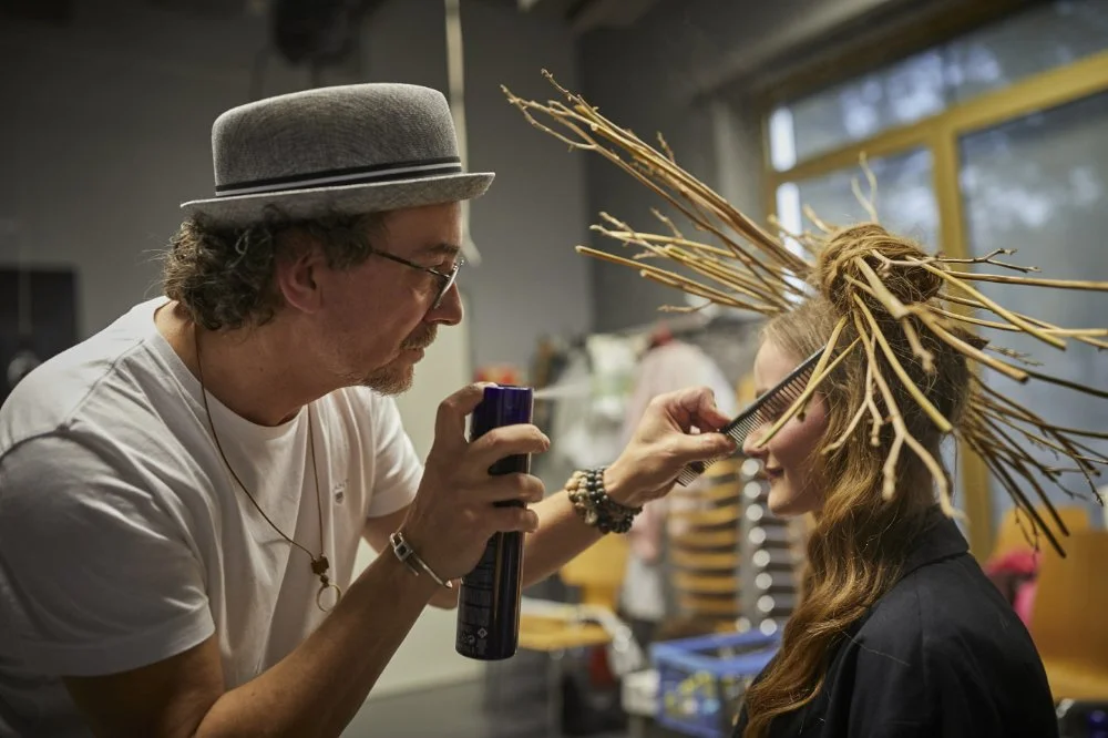 Friseur mit Hut frisiert eine Frau mit einem Haarschmuck aus Zweigen in einem Salon.
