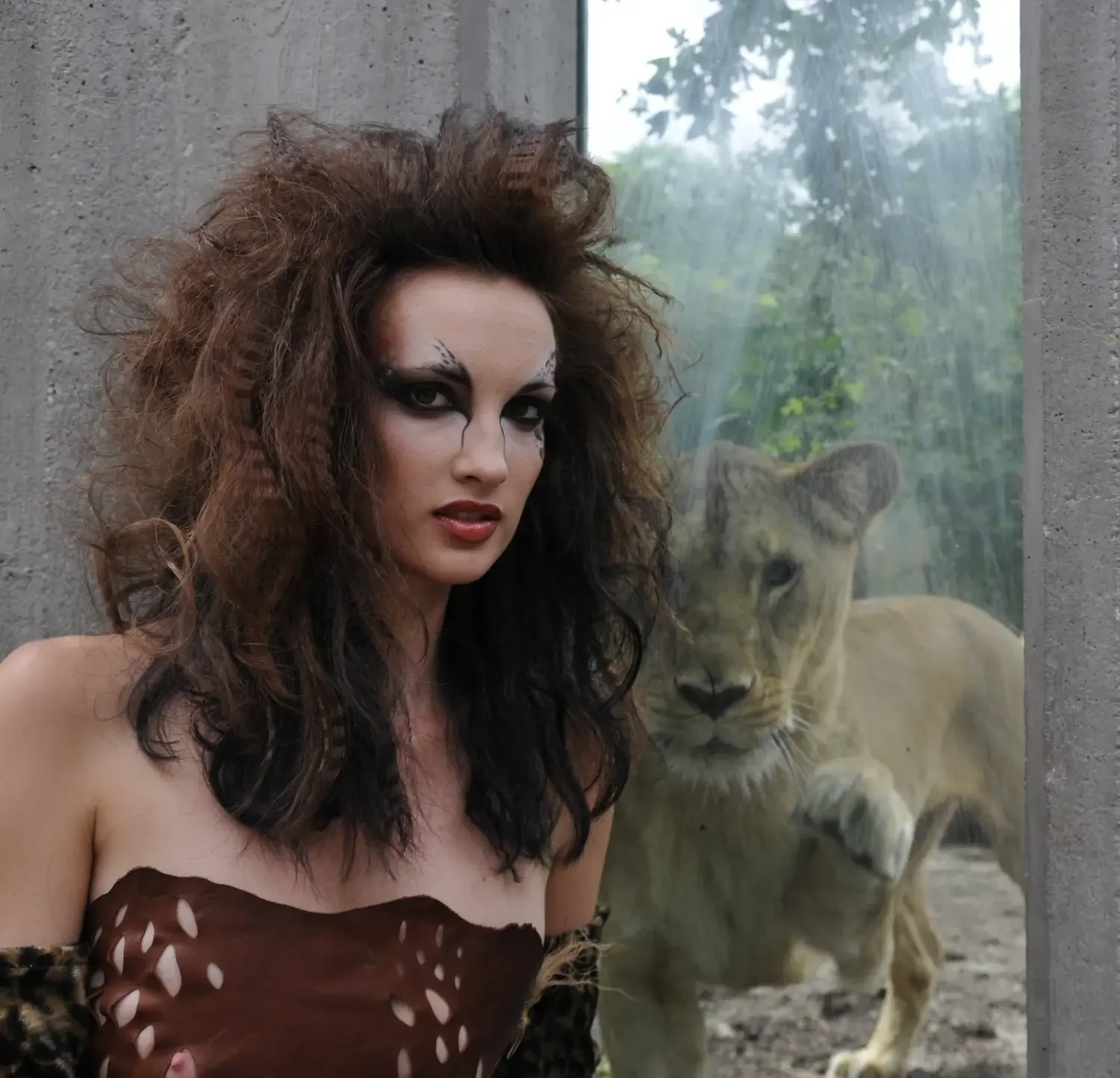 Dramatisches Editorial-Foto mit wildem Hairstyling und intensivem Augen-Make-up vor einer Löwin im Hintergrund – kreative Beauty-Inszenierung im Fashion-Kontext.