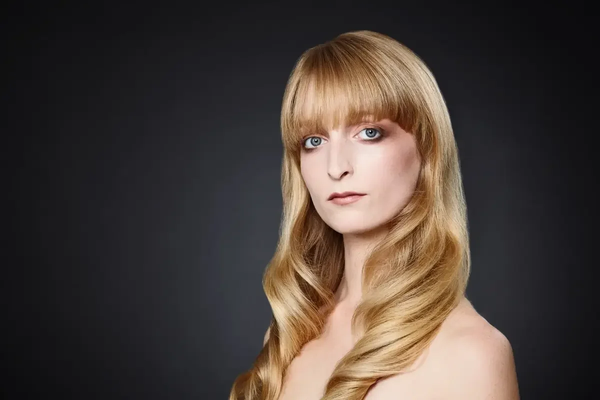 Elegantes Beauty-Porträt mit langen blonden Wellen, geradem Pony und natürlichem Make-up vor dunklem Studiohintergrund. Garzareck Hairstyling mit Fokus auf glänzendes Haar, klare Haut und moderne Editorial-Ästhetik.
