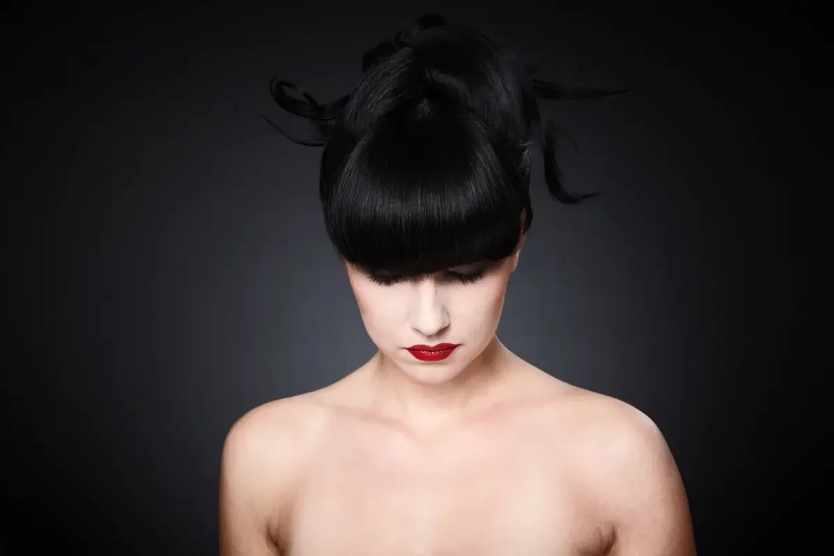 Minimalistisches Beauty-Editorial mit grafischer schwarzer Hochsteckfrisur, porcelainartiger Haut und kräftig roten Lippen vor dunklem Hintergrund. Studioaufnahme mit Fokus auf Haarstyling, Hautbild und moderner Eleganz.