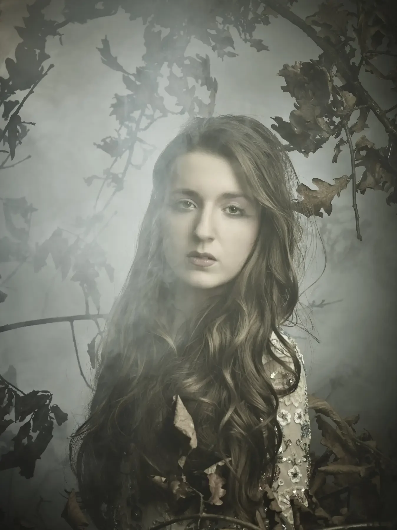 Model mit langen, weichen Wellen und besticktem Kleid vor trockenem Laub und Nebel. Das stimmungsvolle Editorial-Porträt verbindet natürliche Eleganz mit moderner Avantgarde-Ästhetik.