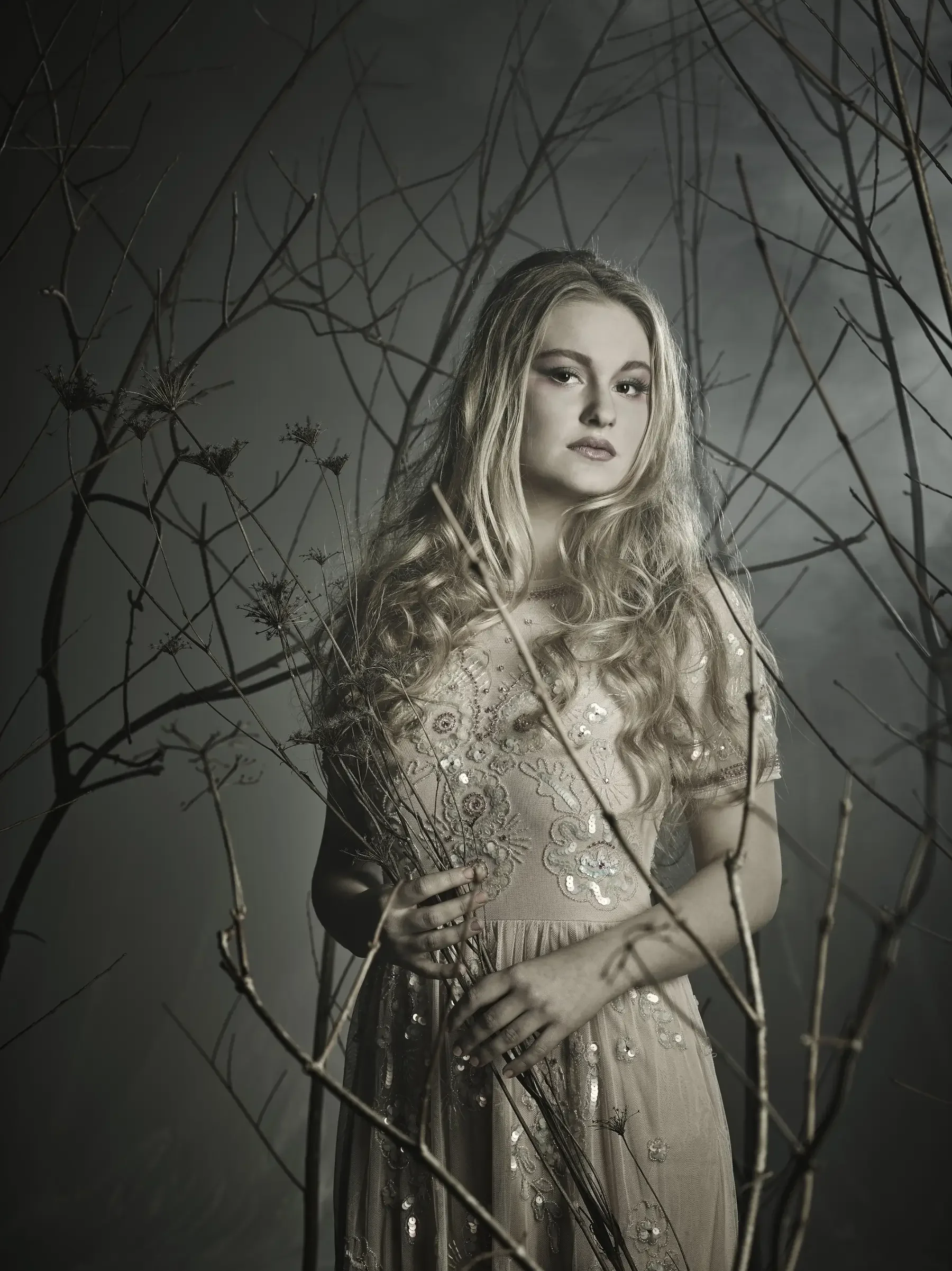 Blondes Model mit weichen Wellen und besticktem Tüllkleid zwischen kahlen Ästen in einer nebeligen Waldszene. Die Editorial-Aufnahme verbindet natürliche Schönheit mit ruhiger, kunstvoller Hair-Fashion-Ästhetik.