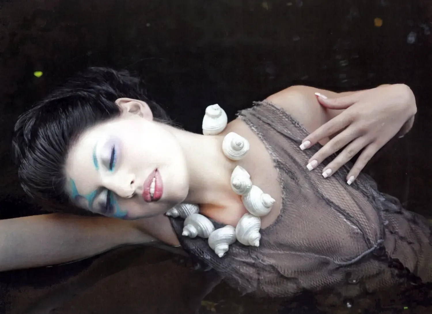 Avantgardistisches Beauty-Porträt mit kreativem Make-up, Muschelschmuck und transparenter Stoffstruktur in dunkler Editorial-Inszenierung.