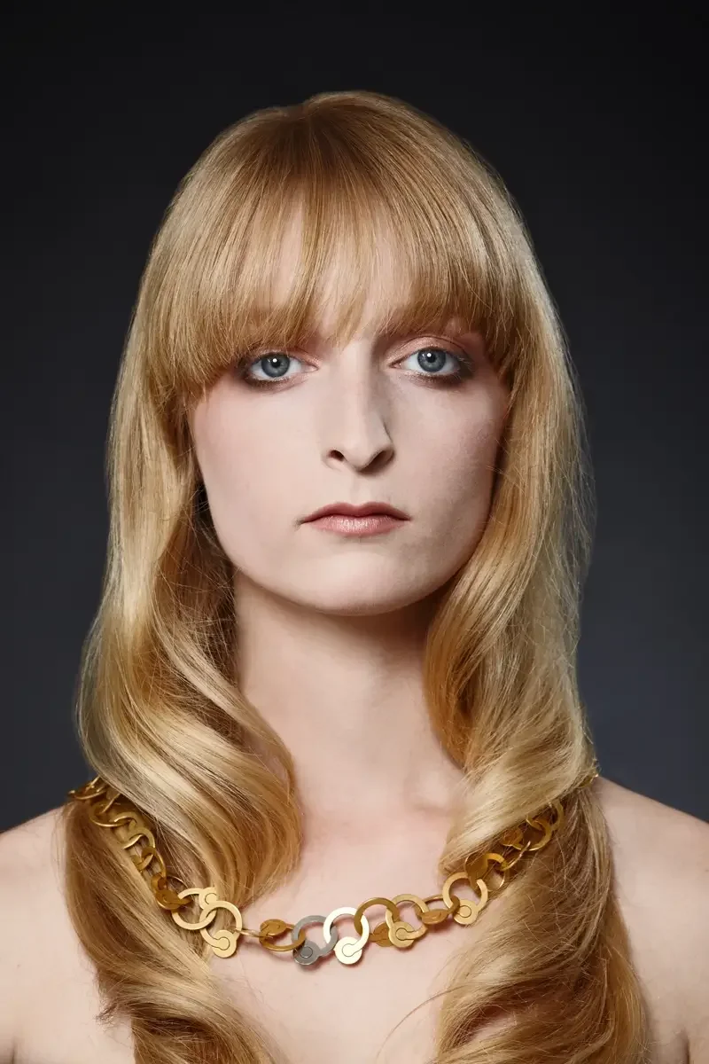 Elegantes Beauty-Porträt mit langen blonden Wellen, geradem Pony und goldfarbenem Designer-Collier vor dunklem Studiohintergrund. Fokus auf glänzendes Haar, klare Haut und hochwertigen Schmuck im modernen Editorial-Stil.