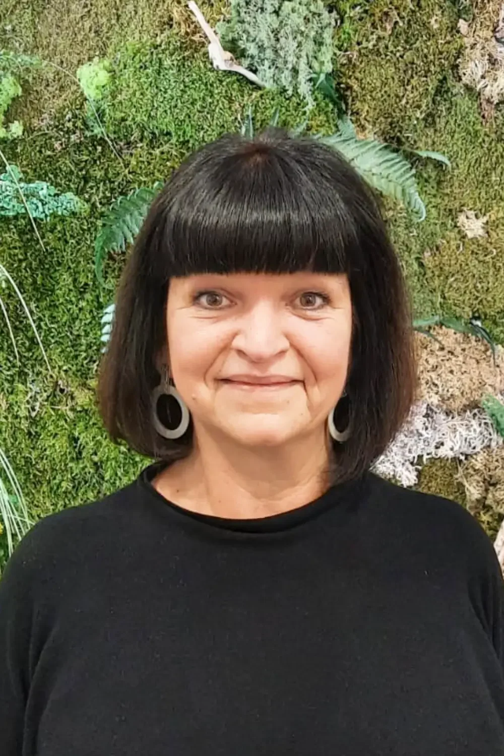 Eine Frau mit schwarzen Haaren und Trichterohrringen, die vor einer grünen Moos- und Pflanzenwand steht.