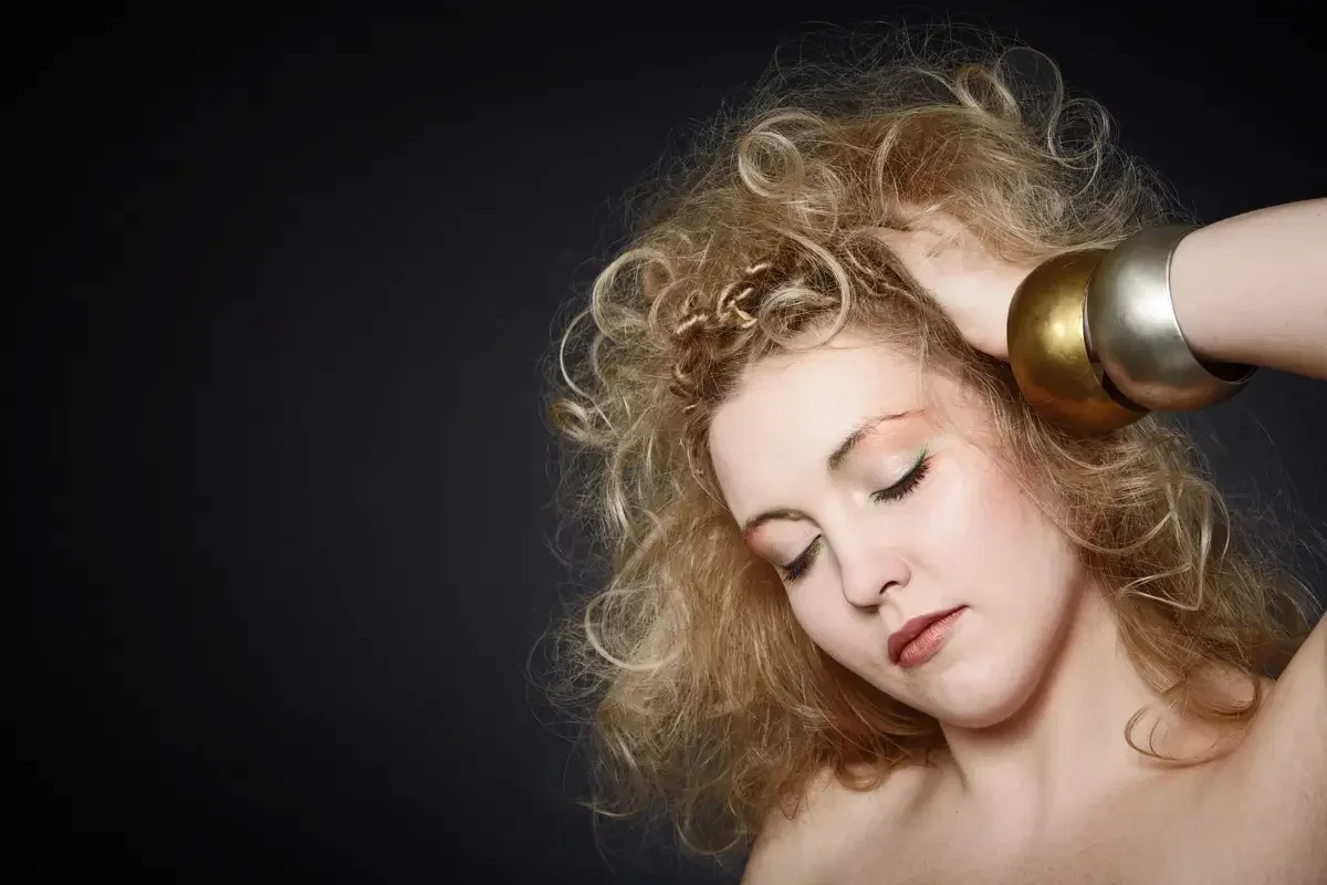 Beauty-Porträt einer Frau mit voluminösen blonden Locken, natürlichem Hautbild und großen goldenen Armreifen vor dunklem Hintergrund. Editorial-Fotografie mit Fokus auf Haut, Haarstruktur, Schmuck und sanftem Make-up.
