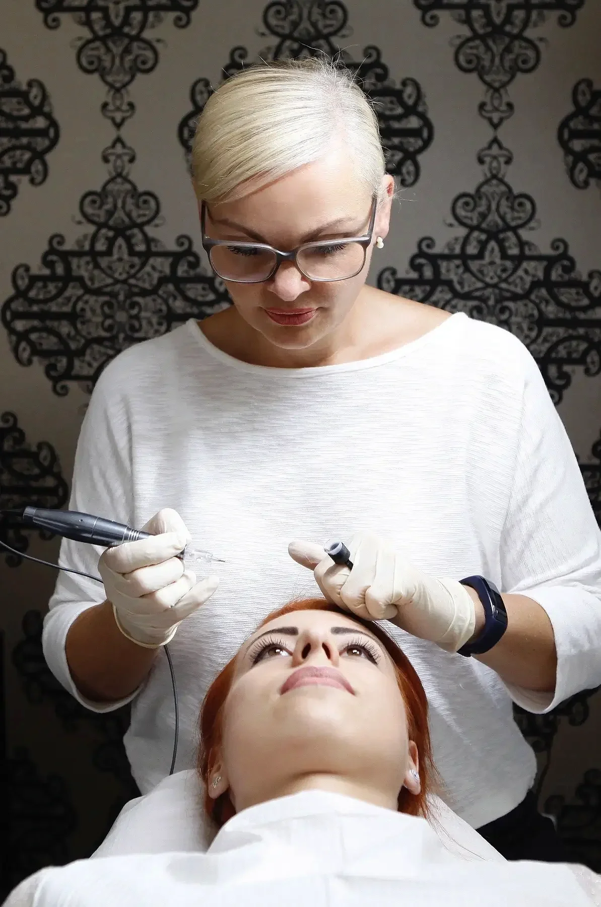 Ariane Garzareck führt eine Permanent-Make-up-Behandlung bei einer Kundin durch, die auf einer Liege liegt. Sie trägt eine Brille, Handschuhe und hält Geräte in den Händen.