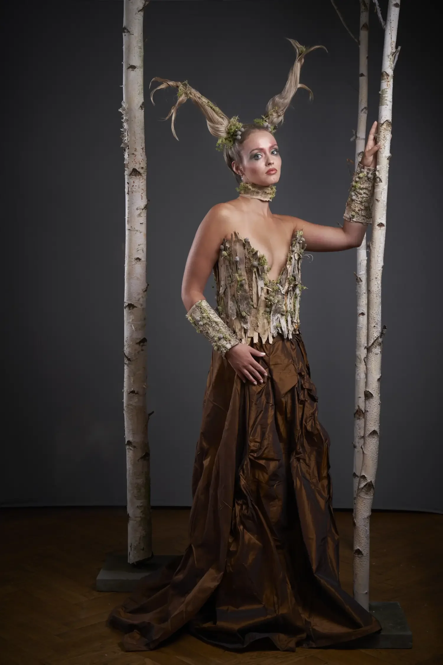 Avantgarde-Modefotografie mit Model im skulpturalen Natur-Couture-Kleid aus Holz, Moos und Rindenstrukturen zwischen Birkenstämmen vor dunklem Studiohintergrund.