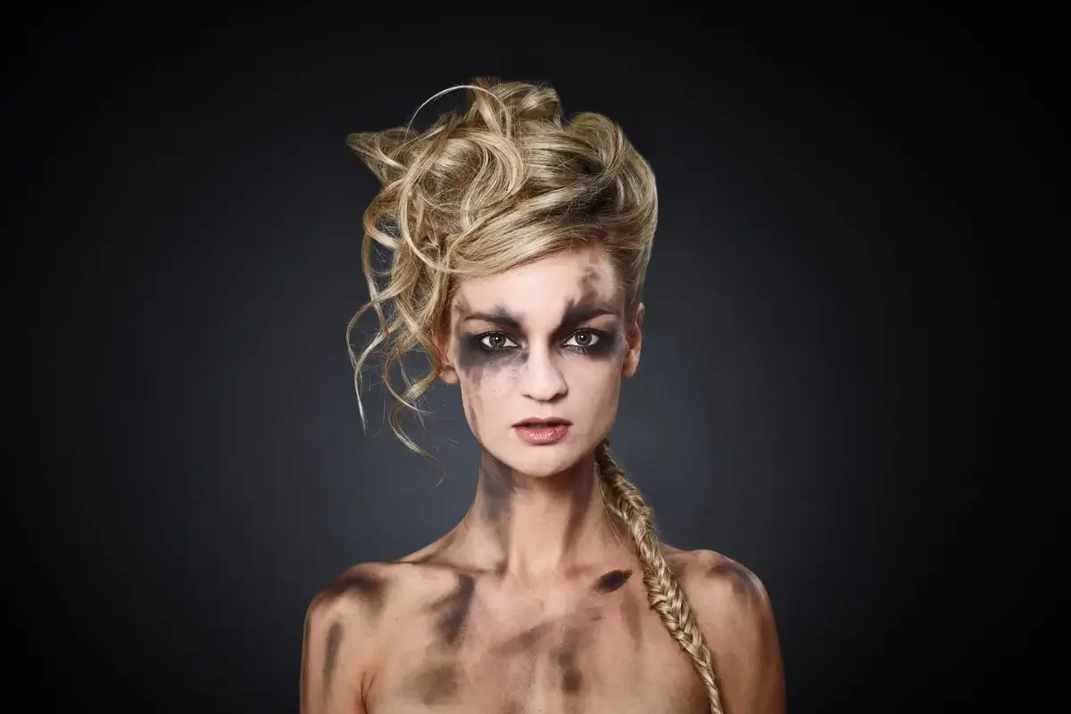 Editorial Beauty-Porträt mit blondem Hochsteckfrisur-Styling, geflochtenem Zopf und dramatischem Smokey-Eye-Make-up vor dunklem Hintergrund. Fokus auf Haut, Haarstruktur und avantgardistische Fashion-Ästhetik.