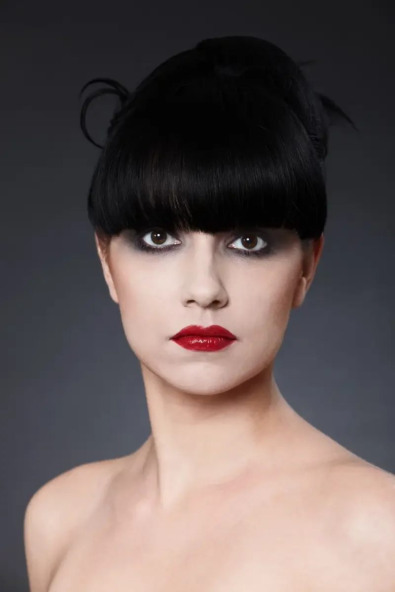 High-Fashion-Beauty-Porträt mit glänzend schwarzer Ponyfrisur, intensiven Smokey Eyes und roten Lippen vor neutralem Studiohintergrund. Editorial-Aufnahme mit Fokus auf Haut, Haarstruktur und modernes Make-up.