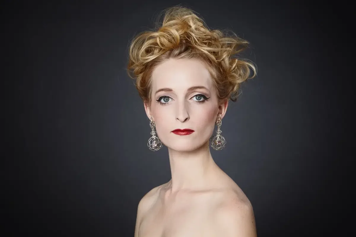Elegantes Beauty-Editorial mit lockiger Hochsteckfrisur, filigranen Statement-Ohrringen und klassischem Make-up vor dunklem Studiohintergrund. Garzareck Inszenierung mit Fokus auf Haut, Haar, Schmuck und zeitloser Schönheit.