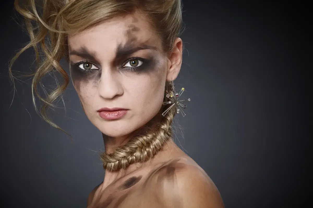 Dramatisches Beauty-Editorial mit verschmiertem Smokey-Eye-Make-up, blondem Avantgarde-Hochsteckfrisur und geflochtenem Haar-Element als Halsaccessoire. Fokus auf Haut, Haar, Schmuck und expressive Fashion-Inszenierung im Studio.