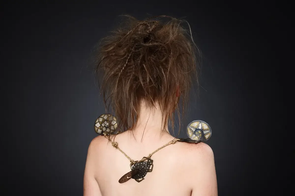 Rückansicht eines avantgardistischen Beauty-Looks mit strukturierter Hochsteckfrisur, handgefertigtem Schmuck und freiem Rücken vor dunklem Studiohintergrund. Künstlerisches Editorial-Styling zu Haut, Haar und Schmuck by Garzareck.