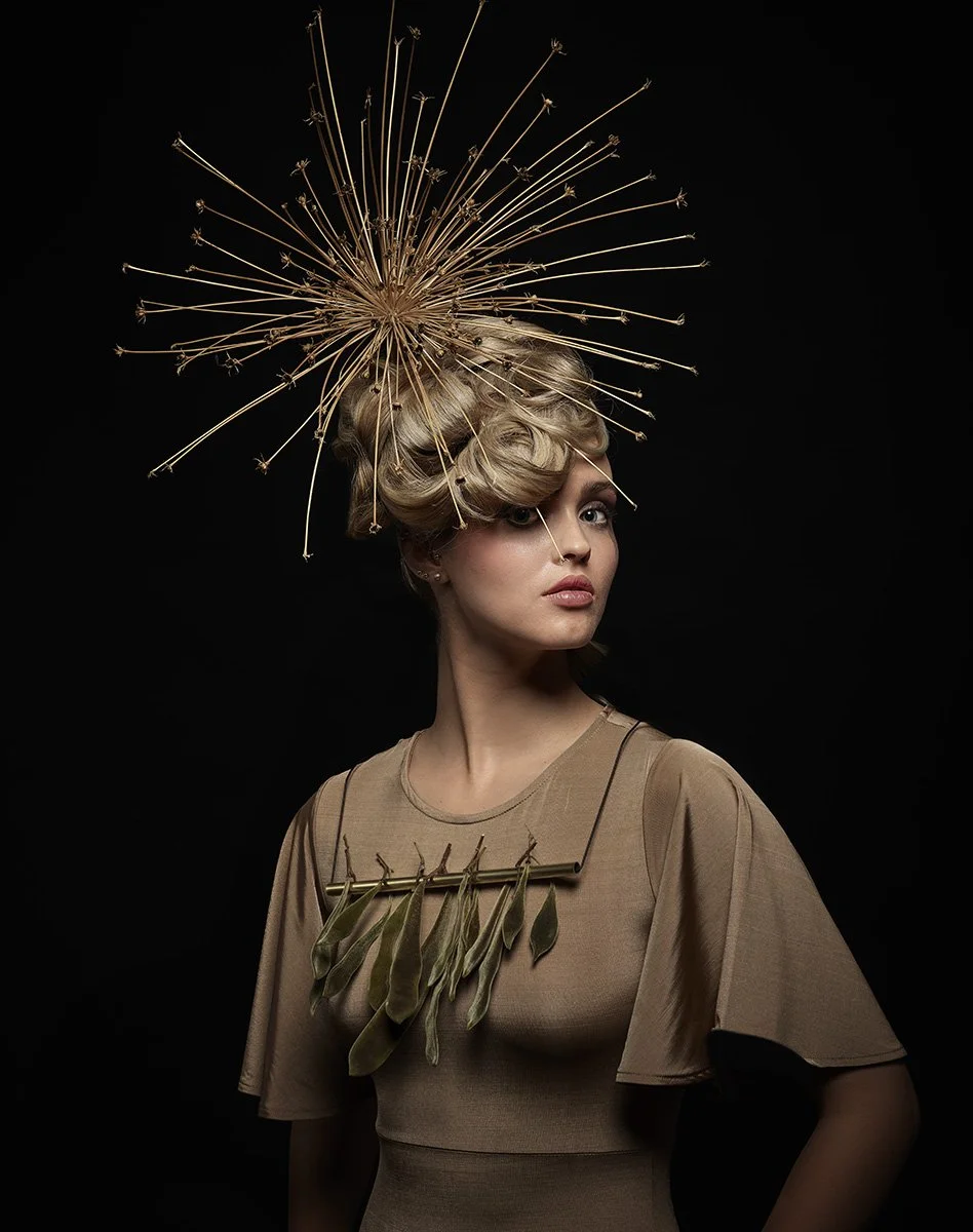 Eine junge Frau mit blond lockigem Haar, die ein Hut aus getrockneten Pflanzen trägt, und ein Kleid mit getrockneten Blättern als Dekoration. Der Hintergrund ist schwarz.