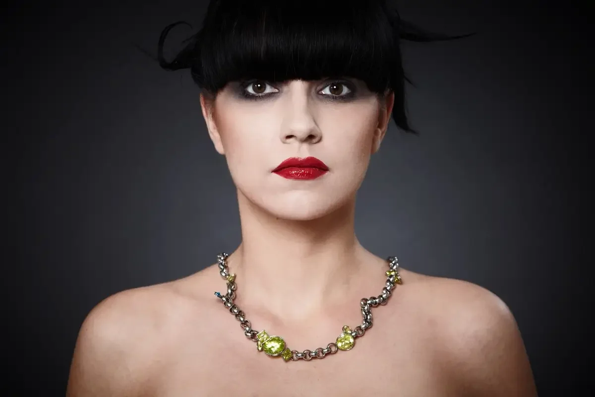 Elegantes Garzareck Beauty-Editorial mit schwarzer grafischer Hochsteckfrisur, markanten roten Lippen und exklusiver Schmuckkette mit grünen Steinen. Studio-Porträt mit Fokus auf Haut, Haar, Schmuck und moderner Luxusästhetik.