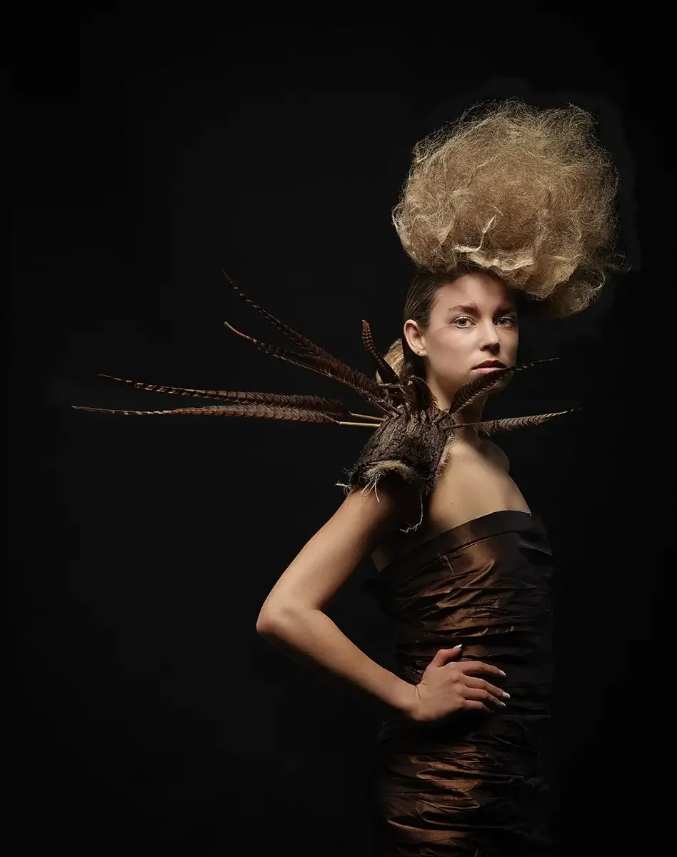 Editorial-Studioporträt mit skulpturaler Hochsteckfrisur, schwarzem Couturekleid und naturinspiriertem Schulterobjekt aus Federn vor dunklem Hintergrund.
