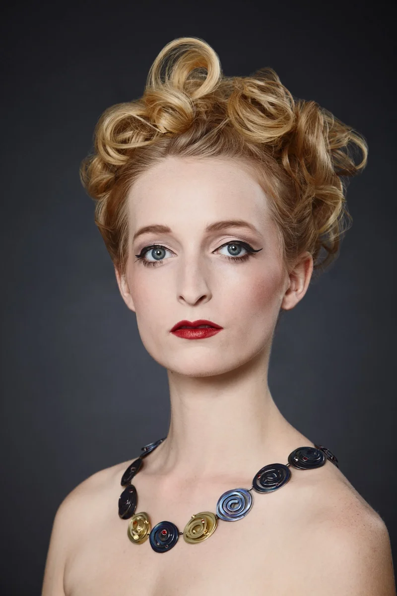 Beauty-Porträt mit kunstvoll gelockter Hochsteckfrisur, rotem Lippenstift und modernem Designer-Collier vor dunklem Studiohintergrund. Fokus auf elegante Haut, Haarstyling und exklusiven Schmuck im Editorial-Look.