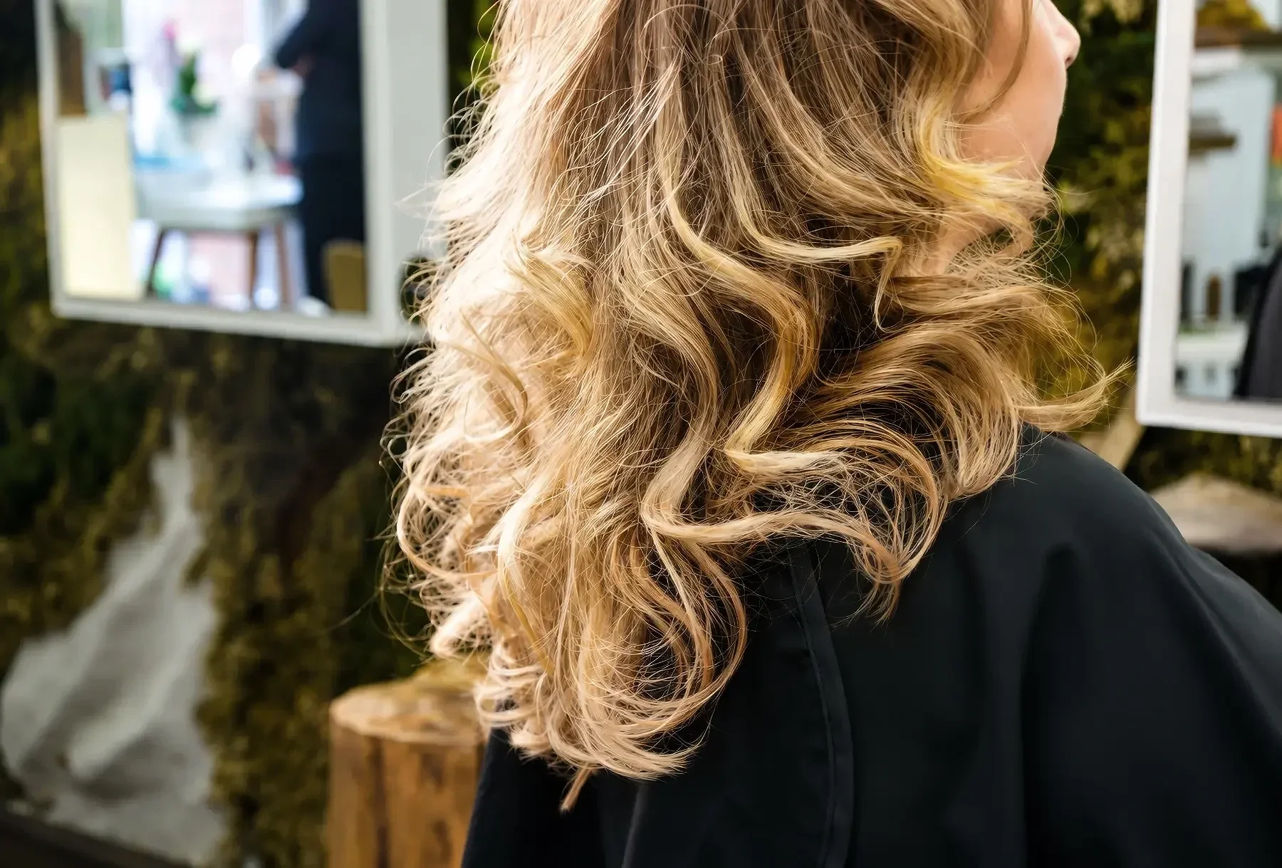 Nahaufnahme von einer Frau mit lockigem, blondem Haar, die seitlich betrachtet wird, vor einem Hintergrund mit grünen Pflanzen und spiegelnden Oberflächen.