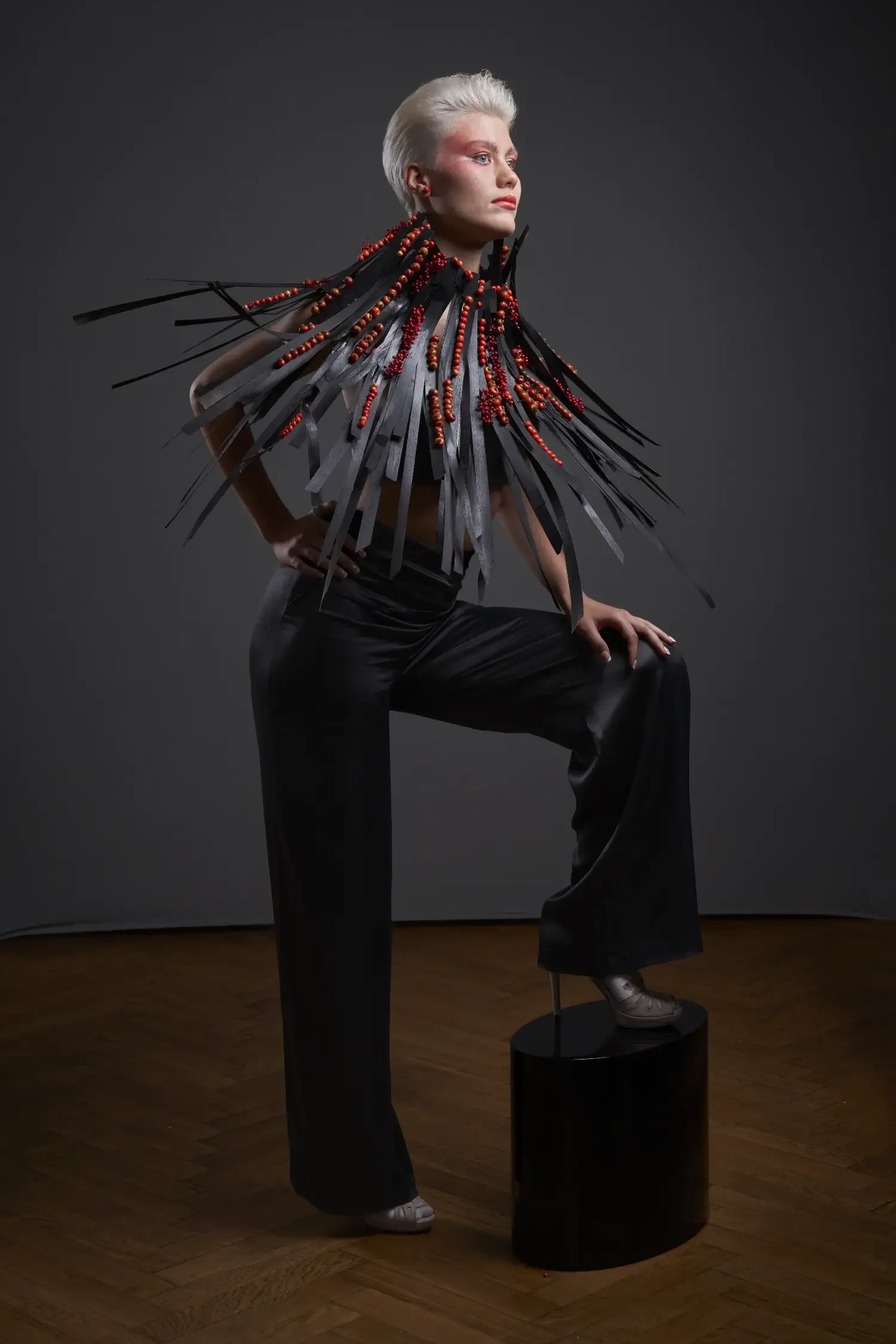 Editorial-Modefotografie mit Model in schwarzer Avantgarde-Couture und skulpturalem Schulterkragen aus schwarzen Elementen und roten Beeren vor dunklem Hintergrund.