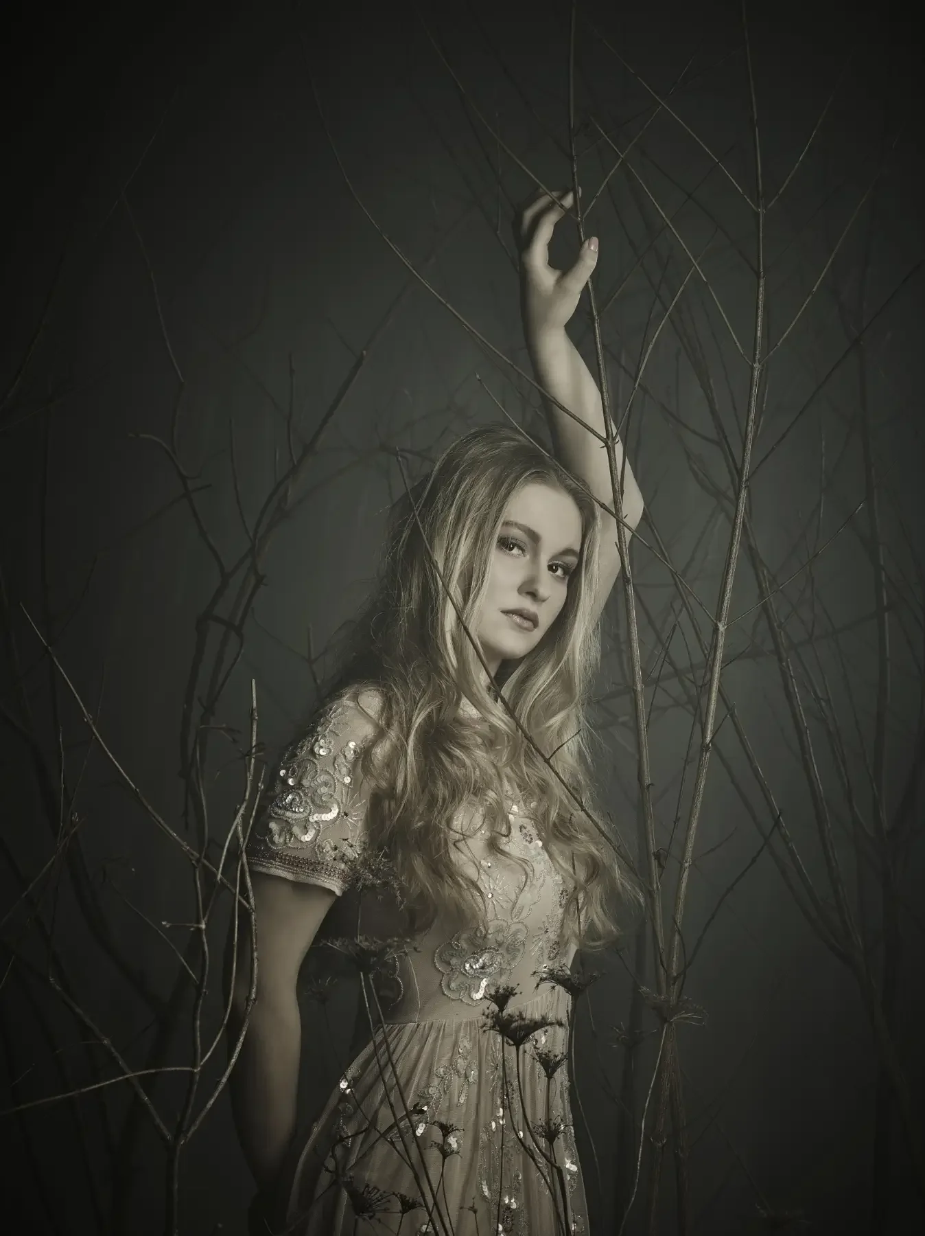 Blondes Model mit weicher Wellenfrisur und besticktem cremefarbenem Kleid zwischen kahlen Ästen. Die dunkle Waldszenerie erzeugt eine ruhige, kunstvolle Editorial-Stimmung.