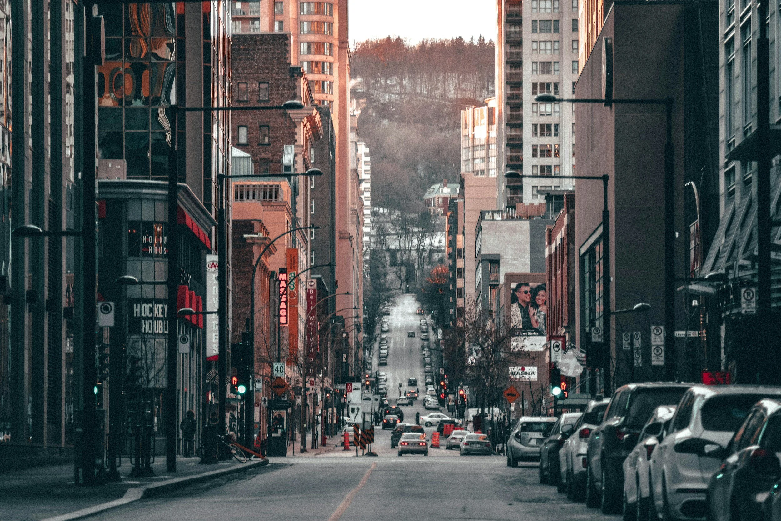 A city street lined with parked cars and tall buildings, with a hill in the background. The street has traffic signals, signs, and advertisements, including a billboard of a man and woman. The scene appears to be during dusk or dawn.