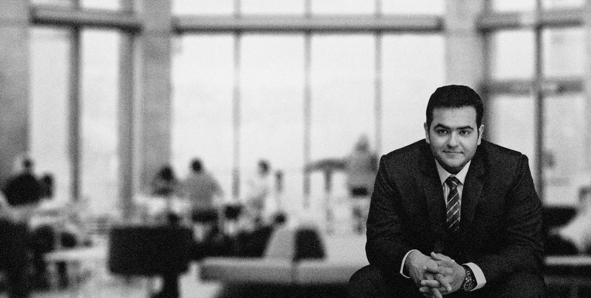 A professional man in a business suit sitting at a table in a modern office with large windows, looking at the camera.