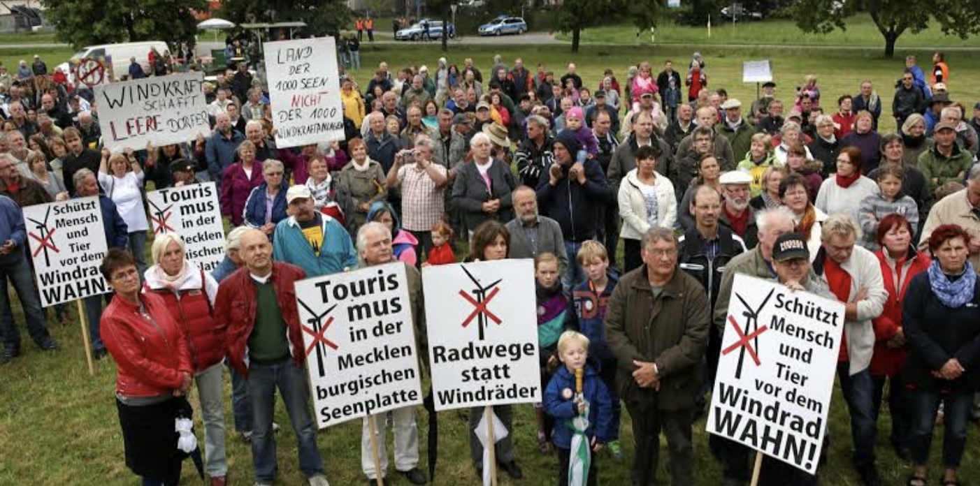 A large gathering of people protesting outdoors, holding signs in German opposing wind turbines and promoting bike paths instead of wind turbines.