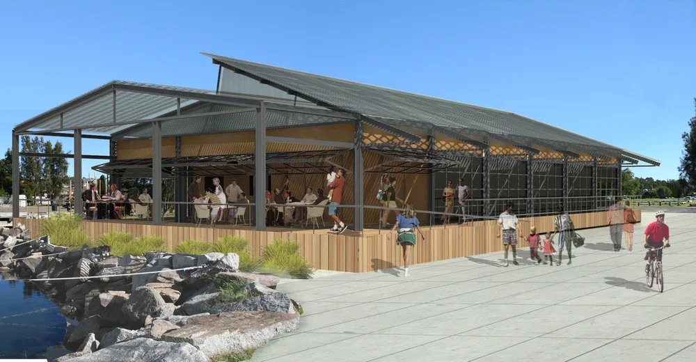 A large open-air community pavilion with a sloped metal roof, situated near a pond with rocks. People are sitting inside at tables, some are walking or cycling outside the pavilion, and a few children and adults are near the entrance.