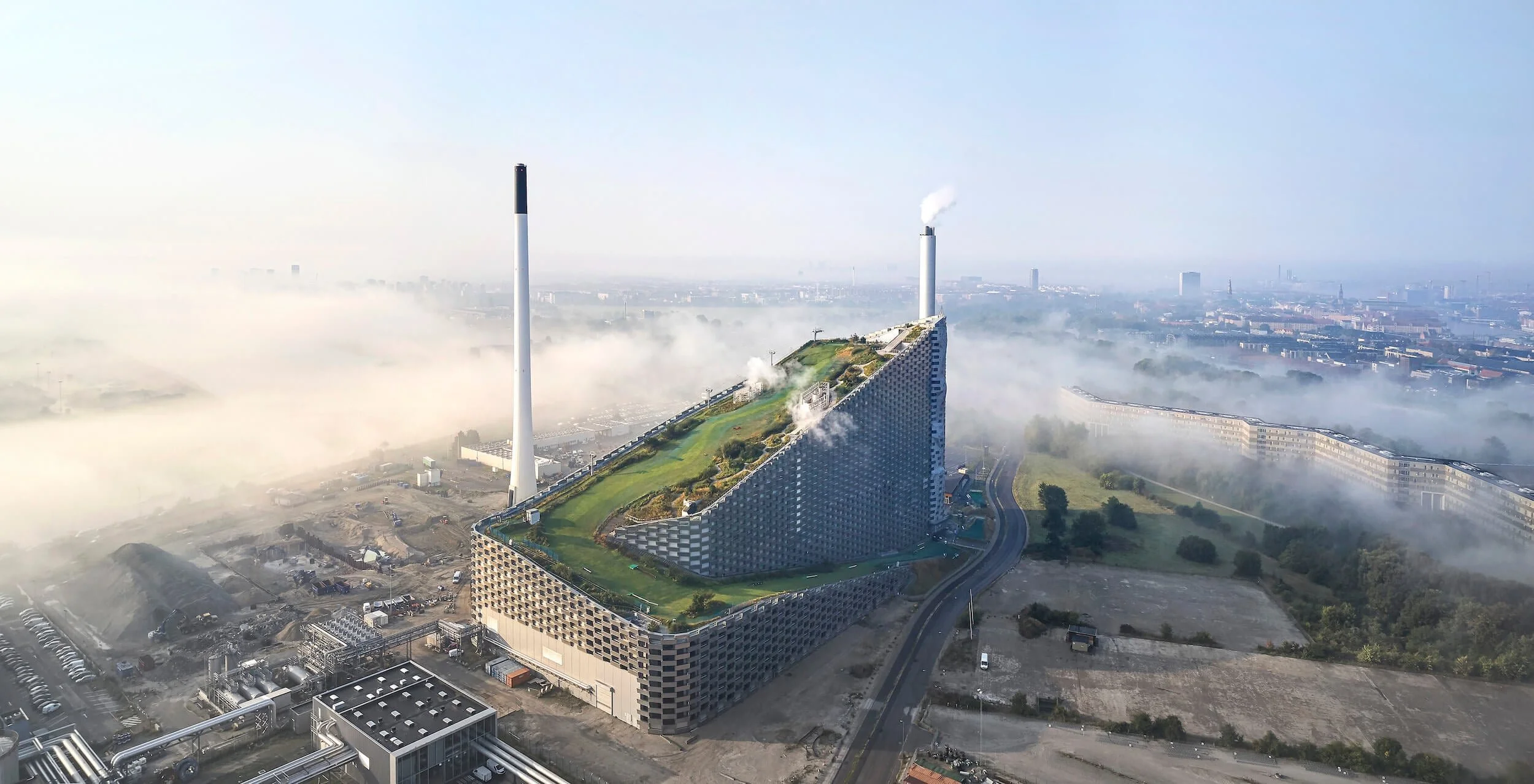 Aerial view of a the trigeneration power plant CopenHill by BIG with a green roof, surrounded by fog, industrial chimneys, and city of Copenhagen skyline in the distance.