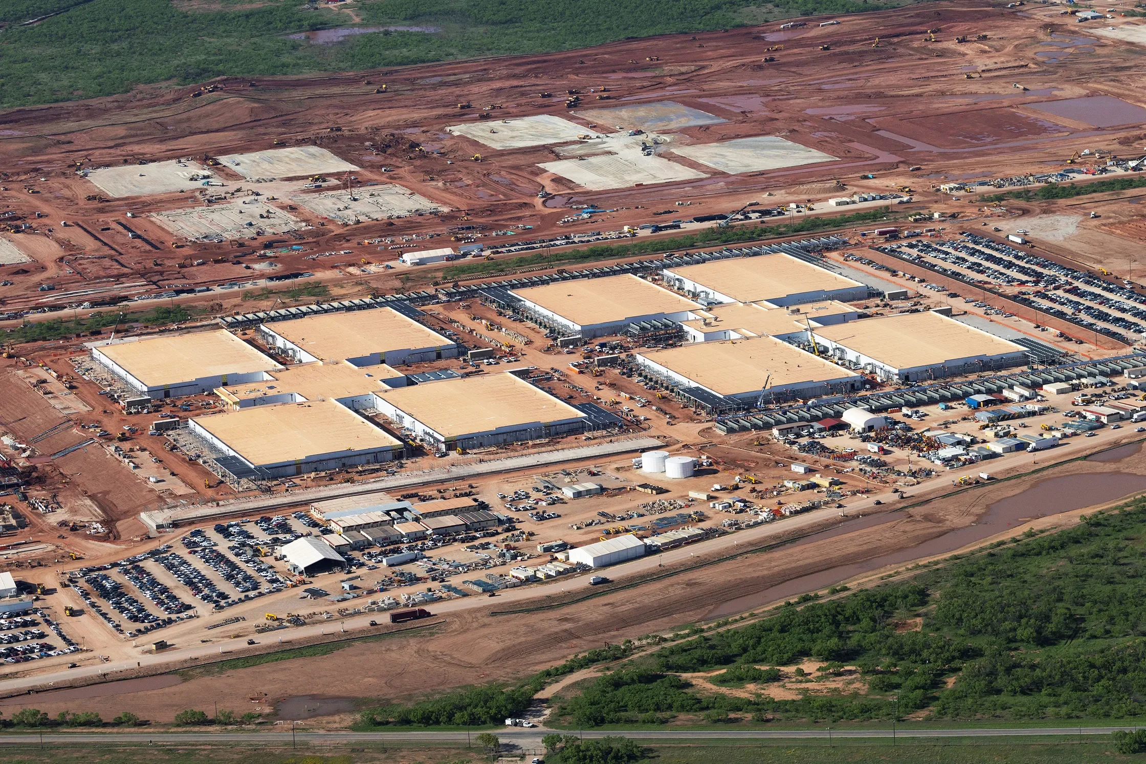 Aerial view of a OpenAI data centre under construction with multiple newly constructed warehouse buildings, parking lots filled with cars, and surrounding land being cleared and prepared for further development.